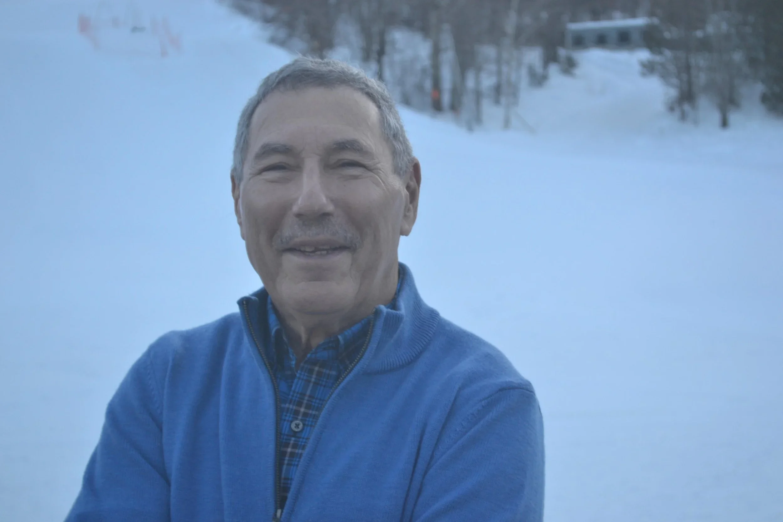 A smiling middle-aged man with gray hair and a mustache taking a selfie outdoors in winter, with snow-covered landscape and trees in the background.