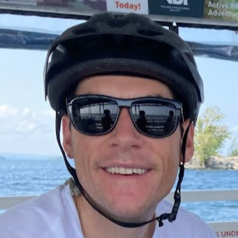 Man wearing a bike helmet and sunglasses, smiling outdoors near water.