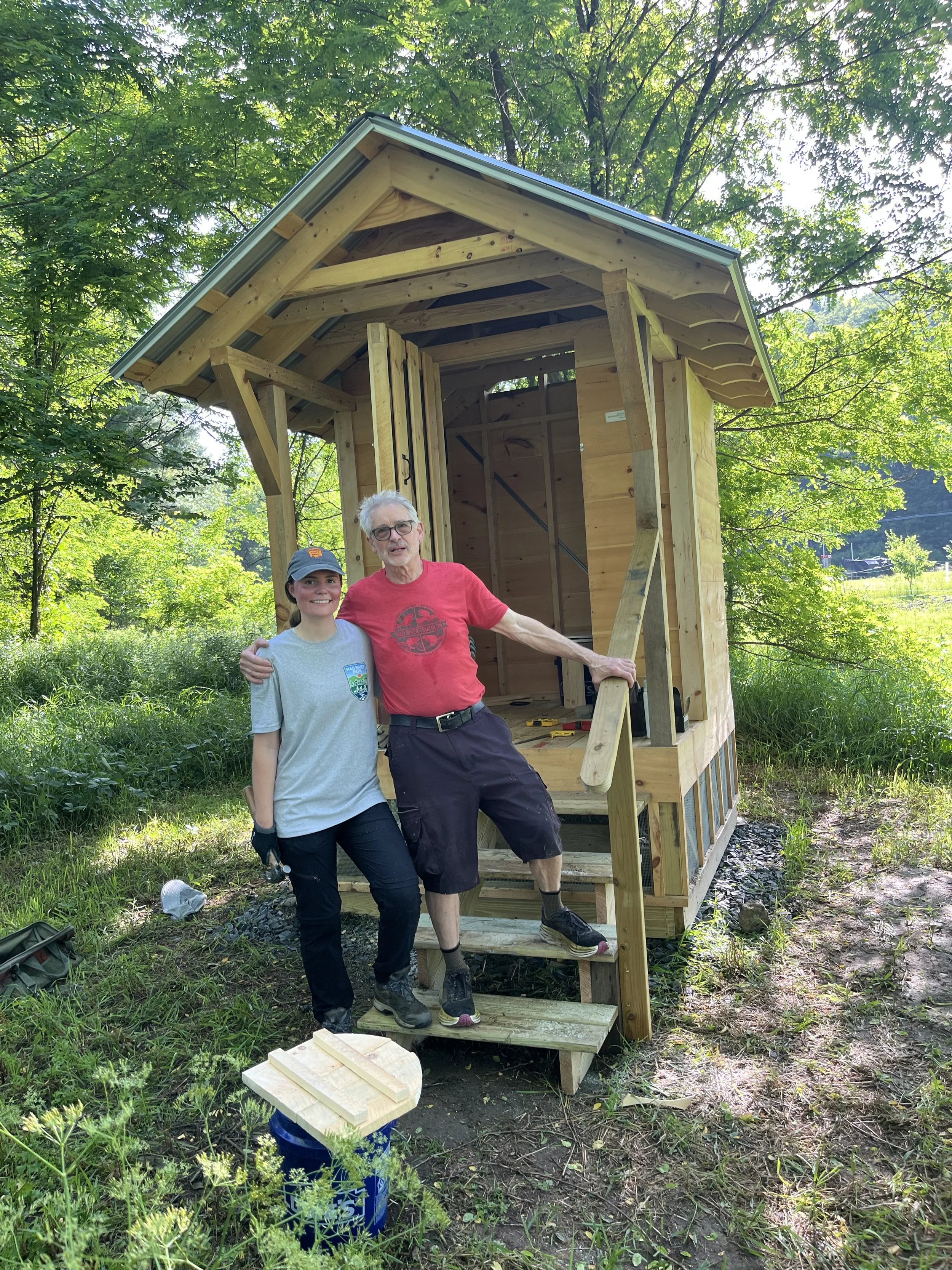 Two people, a young woman and an older man, stand in front of a partially built wooden structure in a green outdoor setting. The structure has a raised platform, ramp, and a small porch with a roof, and they are smiling at the camera.