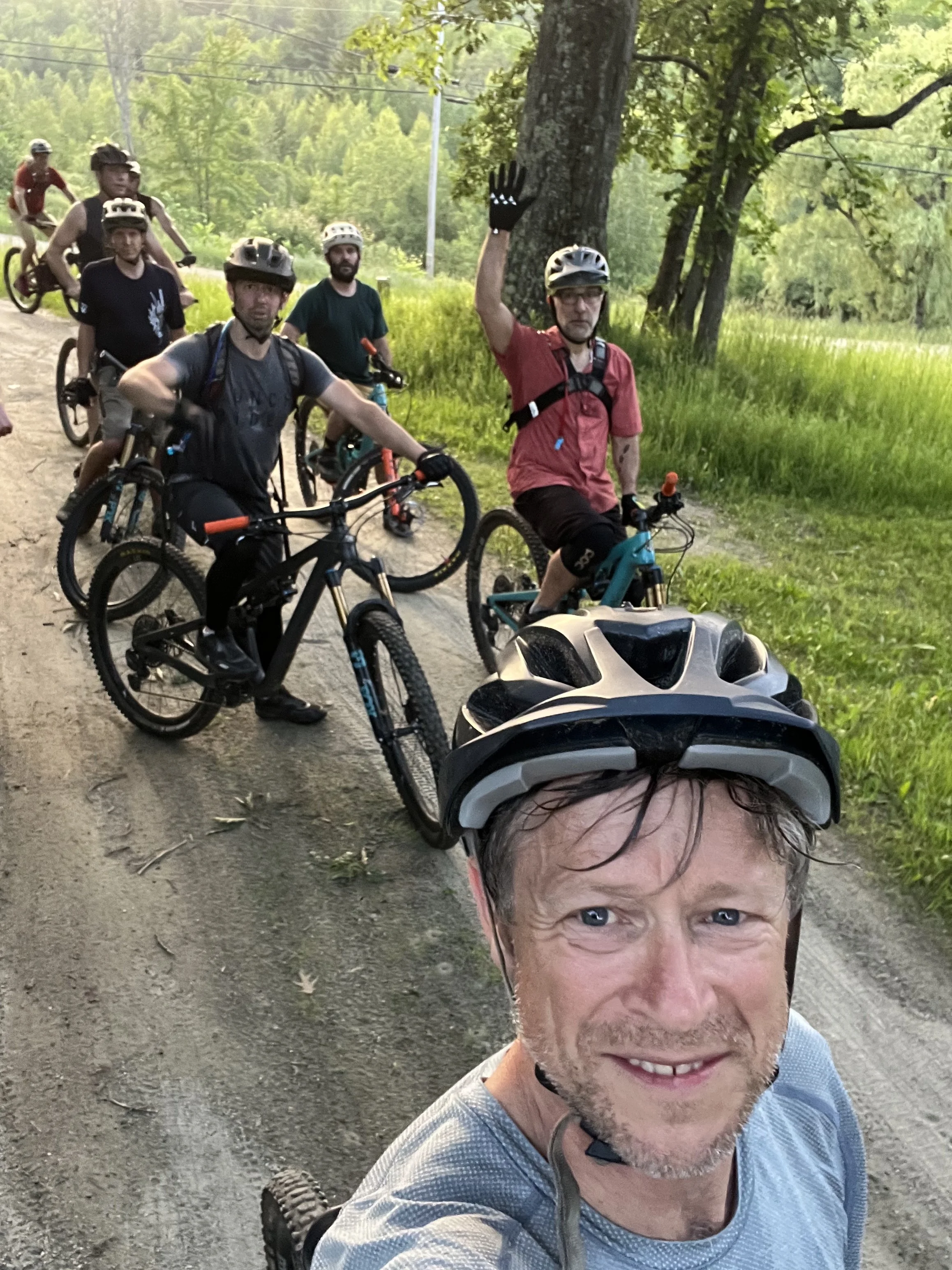 A group of people, mostly men, on mountain bikes on a dirt trail in a green wooded area. One man in the foreground, wearing a helmet and a gray shirt, is taking a selfie and waving at the camera. Other bikers are visible behind him, some wearing helmets and casual clothing, with trees and grass surrounding the trail.