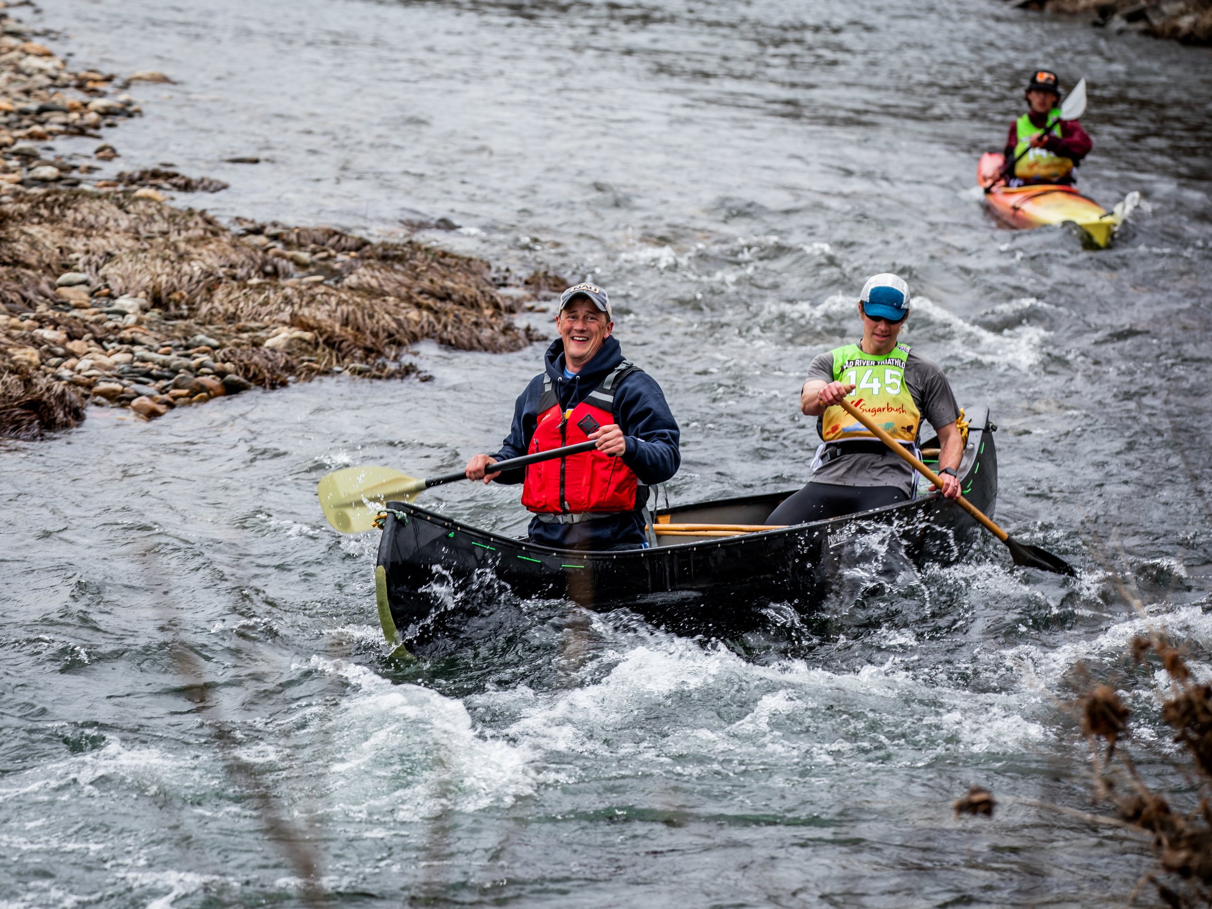 Two men kayaking in a river with rocks and brown algae on the bank, both wearing life jackets and hats, smiling and paddling.
