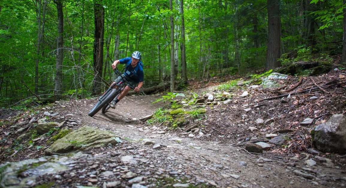 A person mountain biking on a trail through a green forest, leaning into a turn on a dirt path.