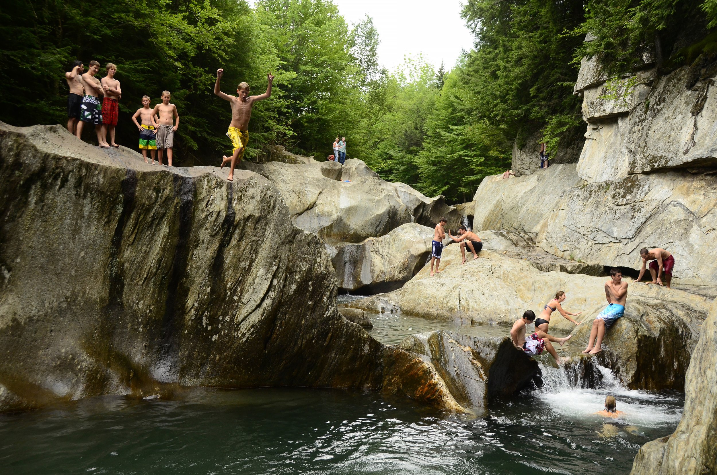 Children and adults swimming and playing on rocks in a river surrounded by lush green trees.