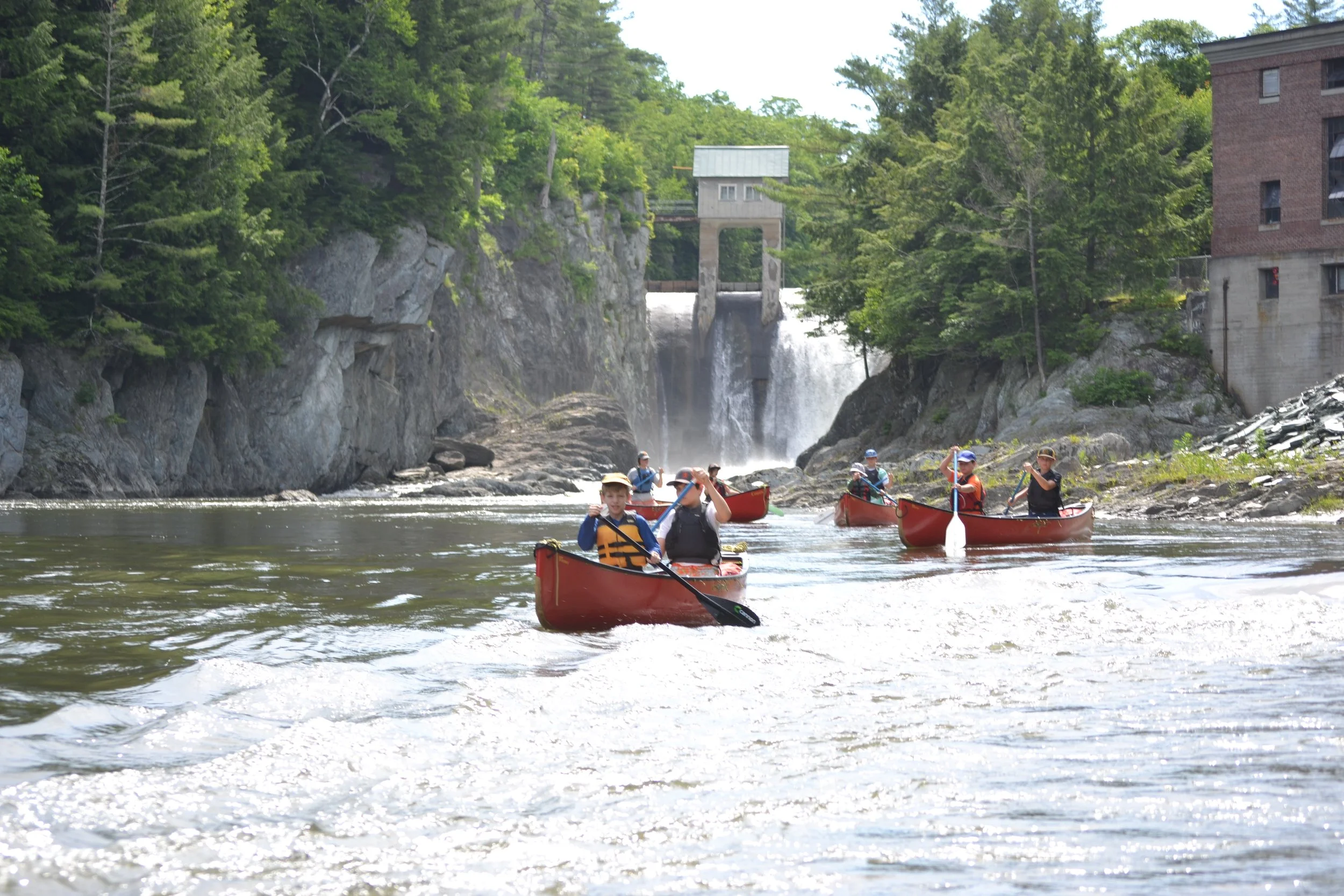 Group of people kayaking on a river near a dam with rocky cliffs and green trees in the background.