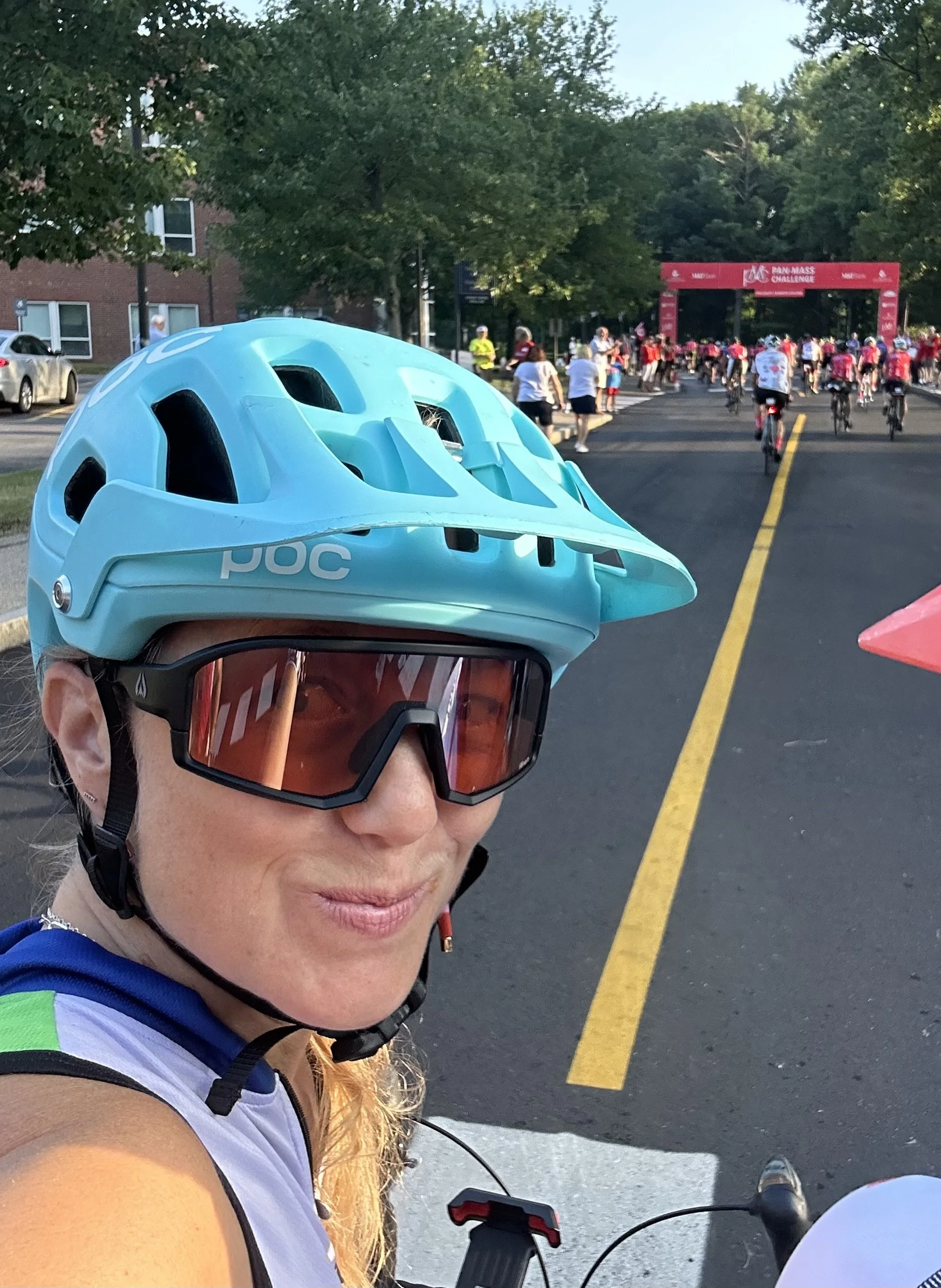 A woman in cycling gear and sunglasses taking a selfie at a cycling event with many cyclists in pink jerseys crossing the finish line in the background.