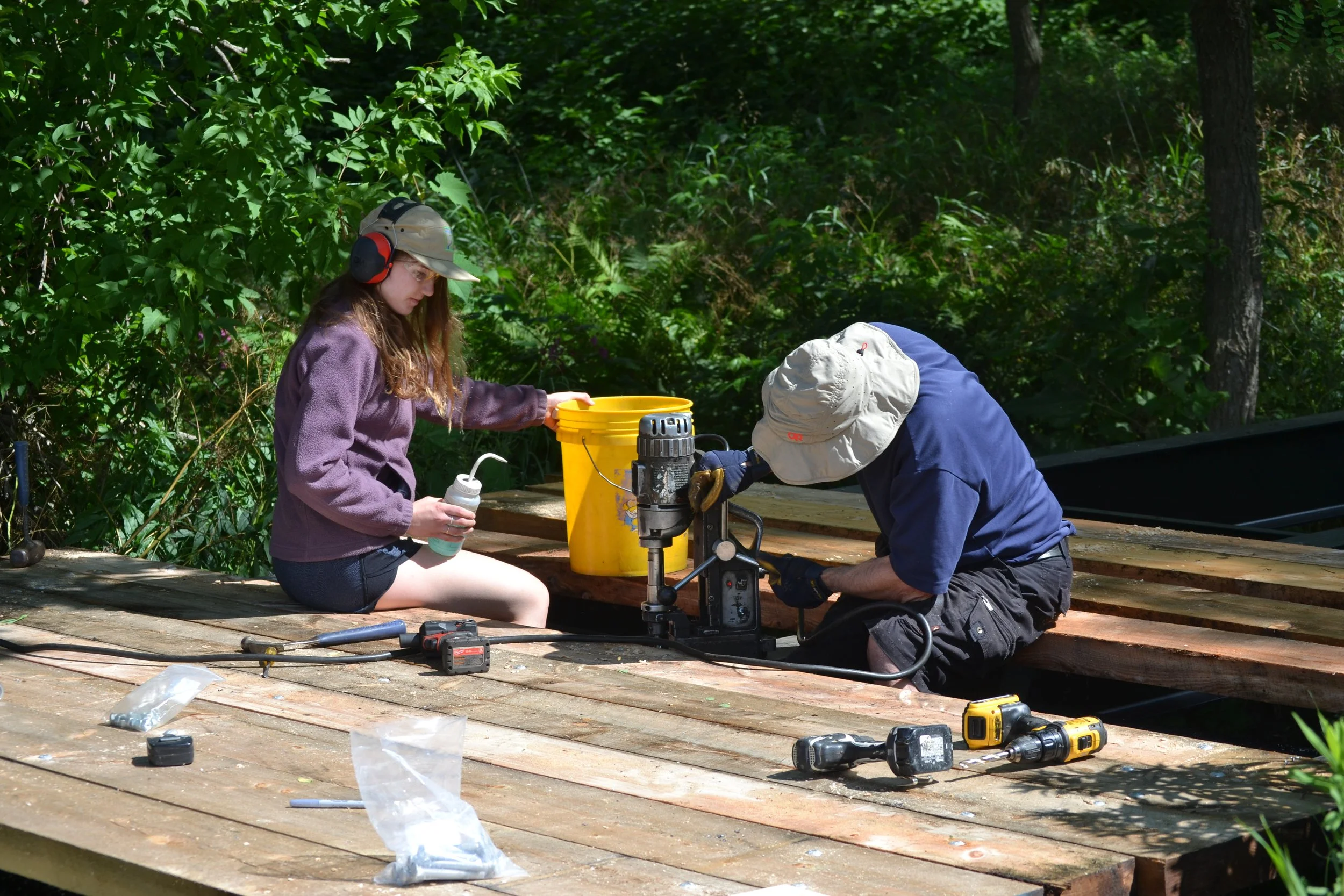Two people working on a construction project outdoors, one young woman and one older man, surrounded by trees and green foliage, using power tools and tools on a wooden surface.