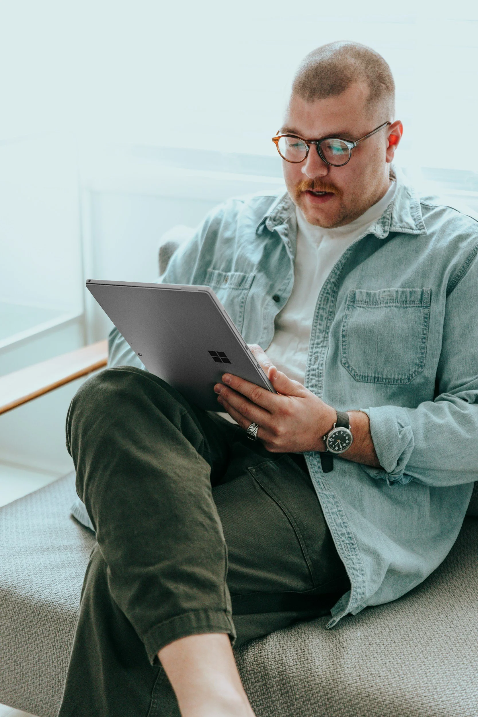 Man met een lichtblauwe jas en een witte shirt zit op een tapijt en kijkt naar een tablet, draagt een horloge en draagt een bril.
