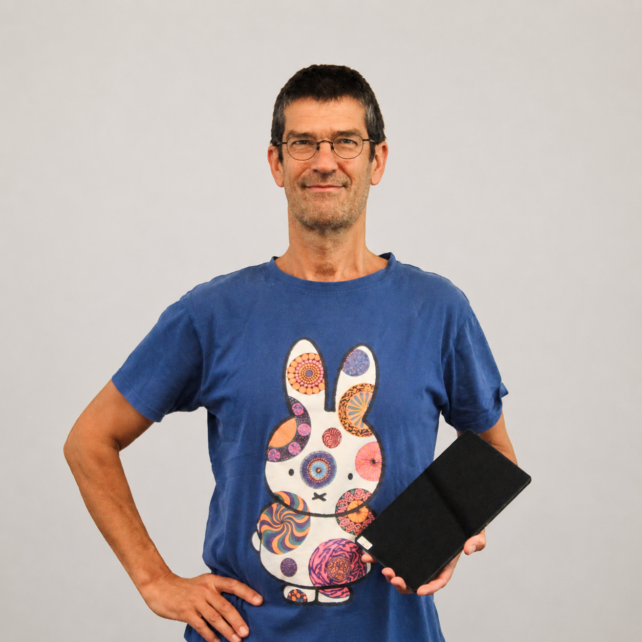 Man met korte donkere haar en brillen, lachend, draagt een blauwe T-shirt met een kleurrijke konijnprint en houdt een zwarte draagbare speaker in zijn rechterhand, staand voor een effen witte achtergrond.