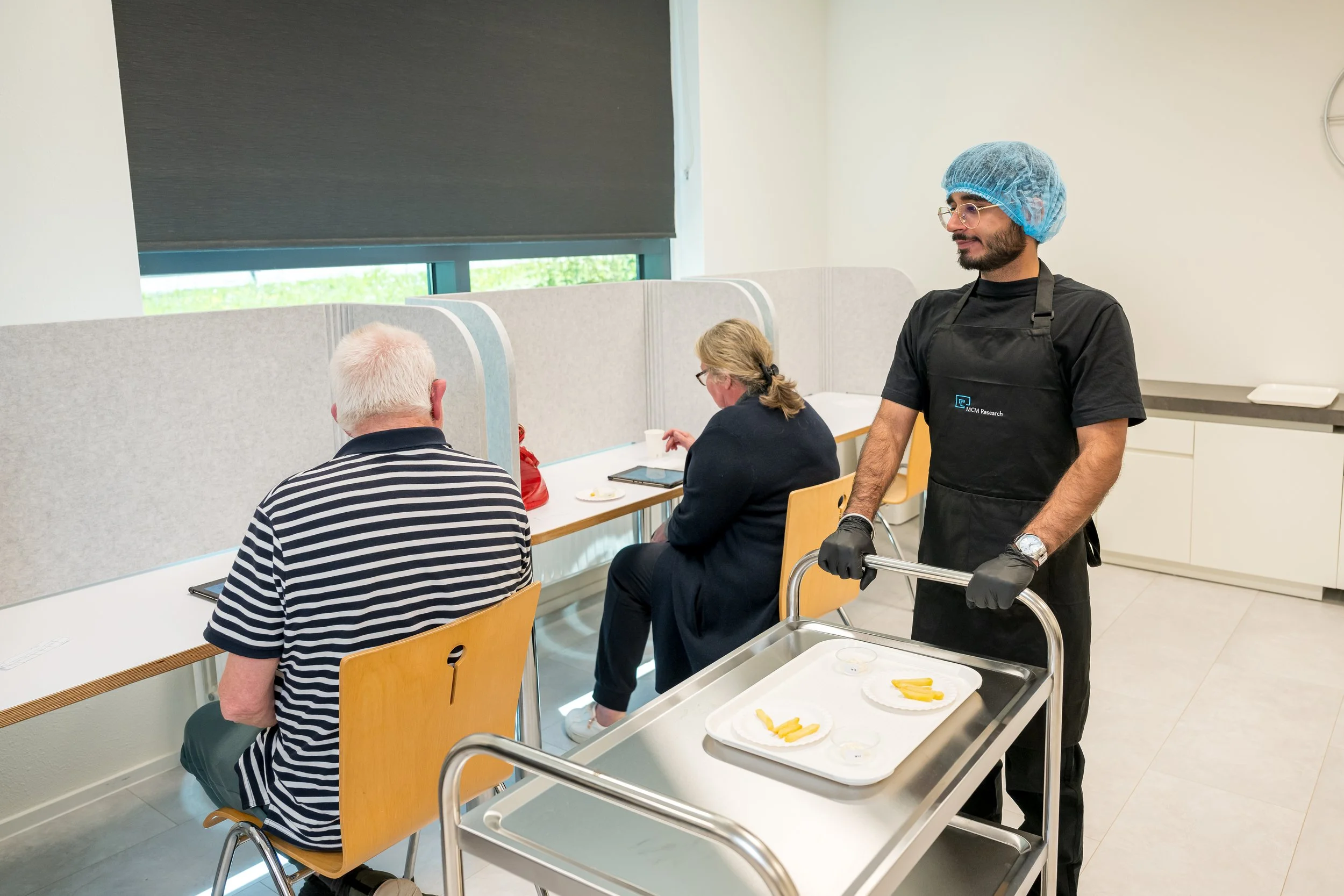 Een man draagt een zwart uniform en een blauw haarnetje, en staat naast een karretje met eten. Hij zorgt voor twee patiënten die aan een tafel zitten, gescheiden door scheidingswanden.