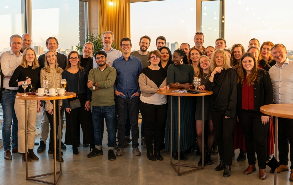 Een grote groep mensen poseert in een zonnig kantoor met uitzicht op de stad door grote ramen.