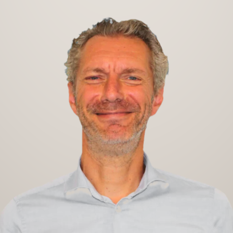 Portret van een lachende man met grijs haar en een witte blouse, voor een neutrale achtergrond.