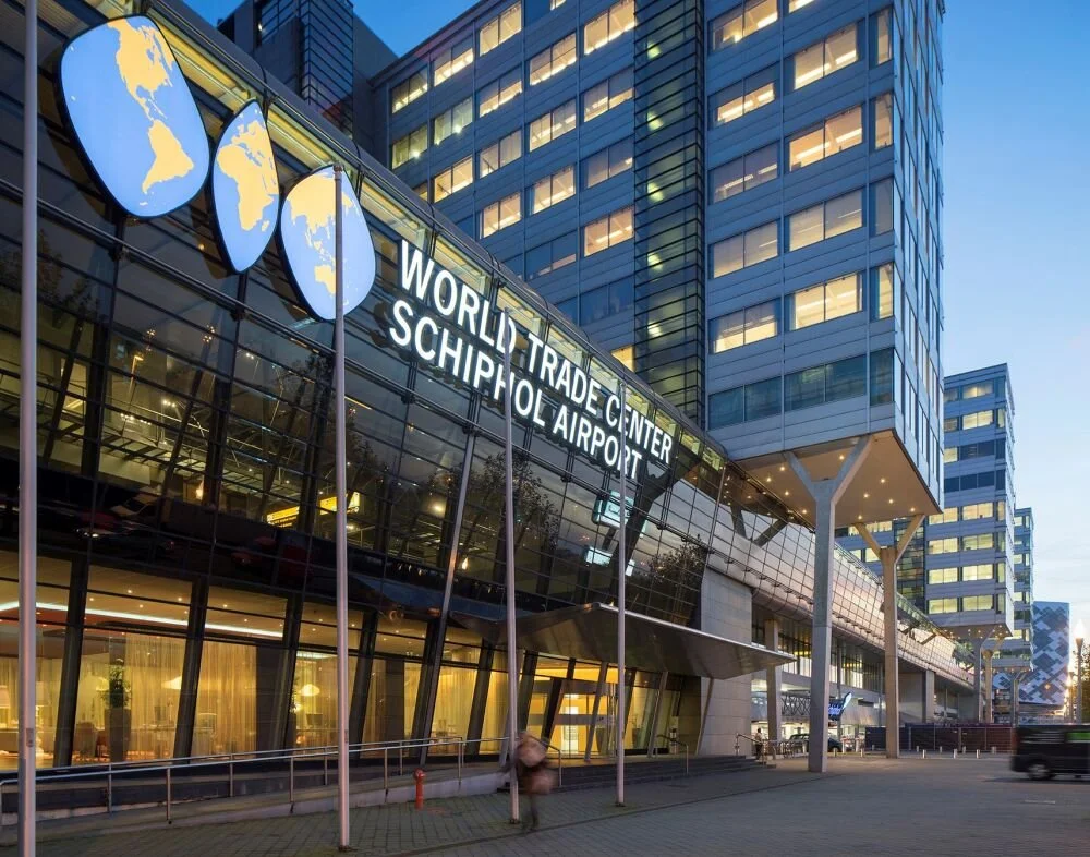 Modern gebouw met een grote glas gevel, met borden die de Wereldhandelscentrum en Schiphol Airport aangeven, en wereldbol-afbeeldingen.