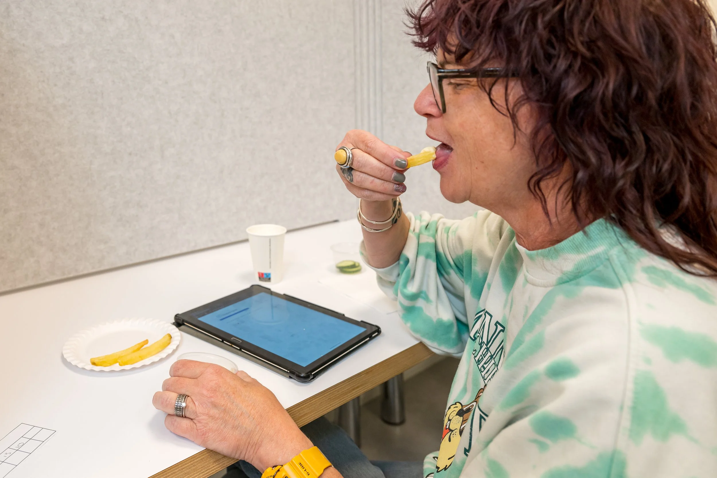 Vrouw met bruin krullend haar, zwarte bril, medaille op haar rechter pols, geel horloge, zittend aan een tafel, eet friet uit haar hand, naast een tablet en een papiertje, op de achtergrond een lichtgrijze muur.