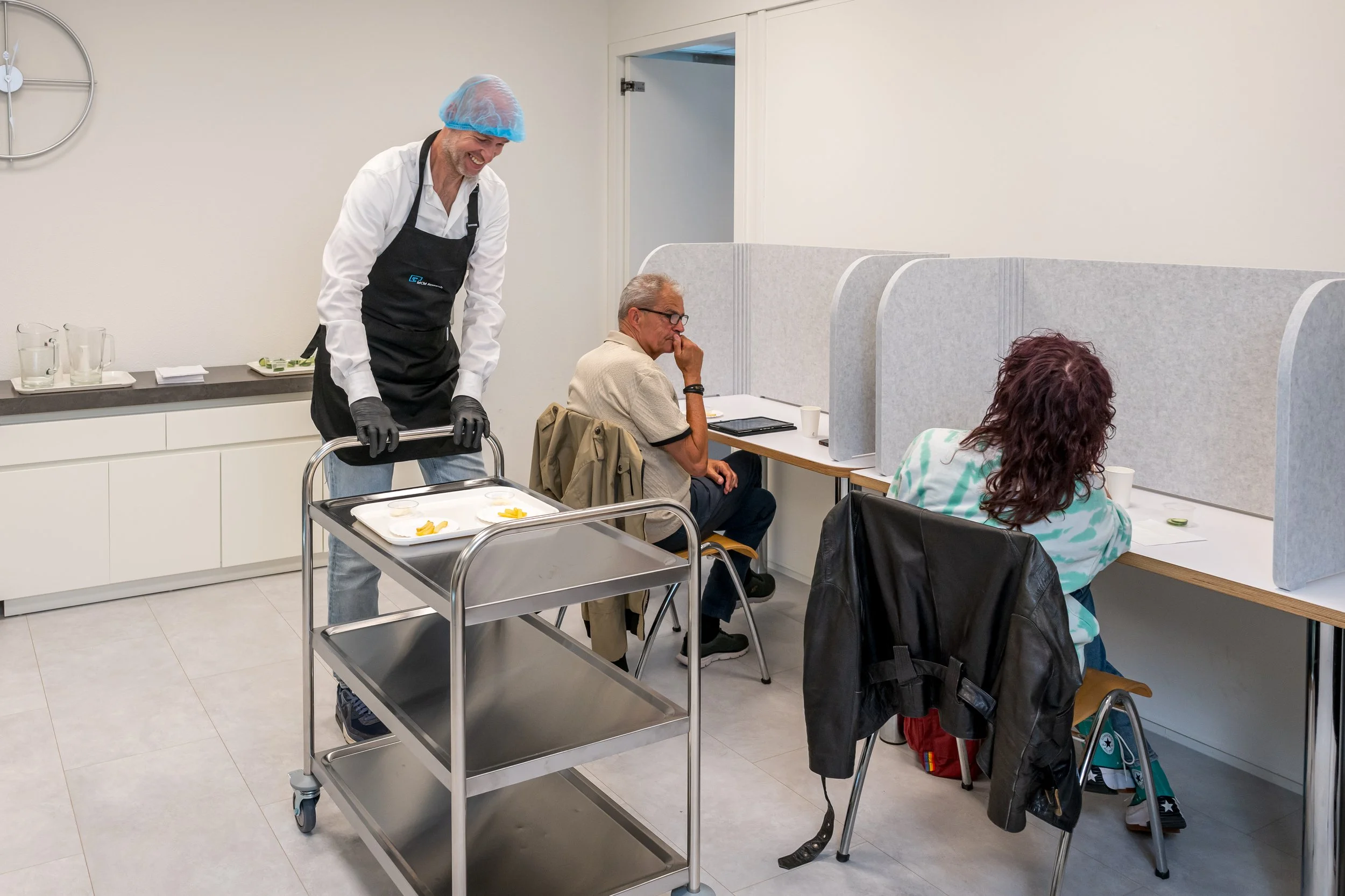 Persone die in een gezondheidskliniek wachten, gescheiden door scheidingswanden, terwijl ze zitten op stoelen met een arts of medisch personeel die door de ruimte loopt.