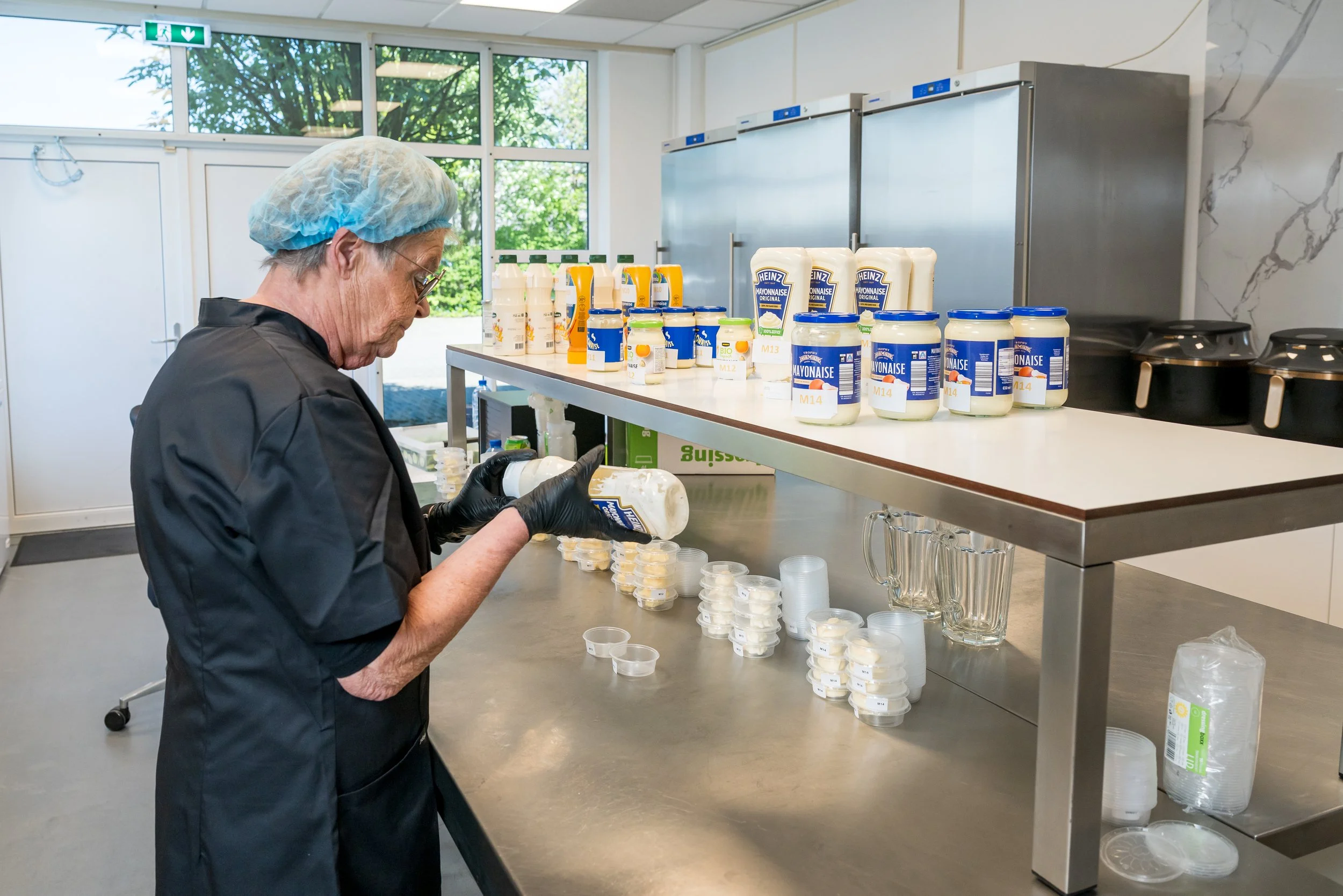 Een persoon met een blauwe haarkap en zwarte handschoenen die smallere containertjes vult met een witte saus, mogelijk mayonaise, in een keuken met producten en containers op de tafel.