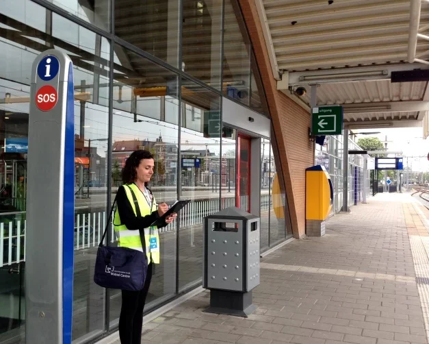 Vrouw met een hoge zichtbare veiligheidsvest en tas wacht naast een informatiepaal met SOS-knop op een treinplatform.