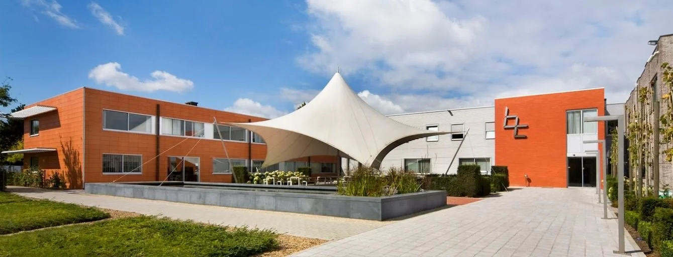 Modern gebouw met een grote witte tentachtige structuur in het midden, omgeven door groen en een betegeld plein onder een blauwe lucht met enkele wolken.