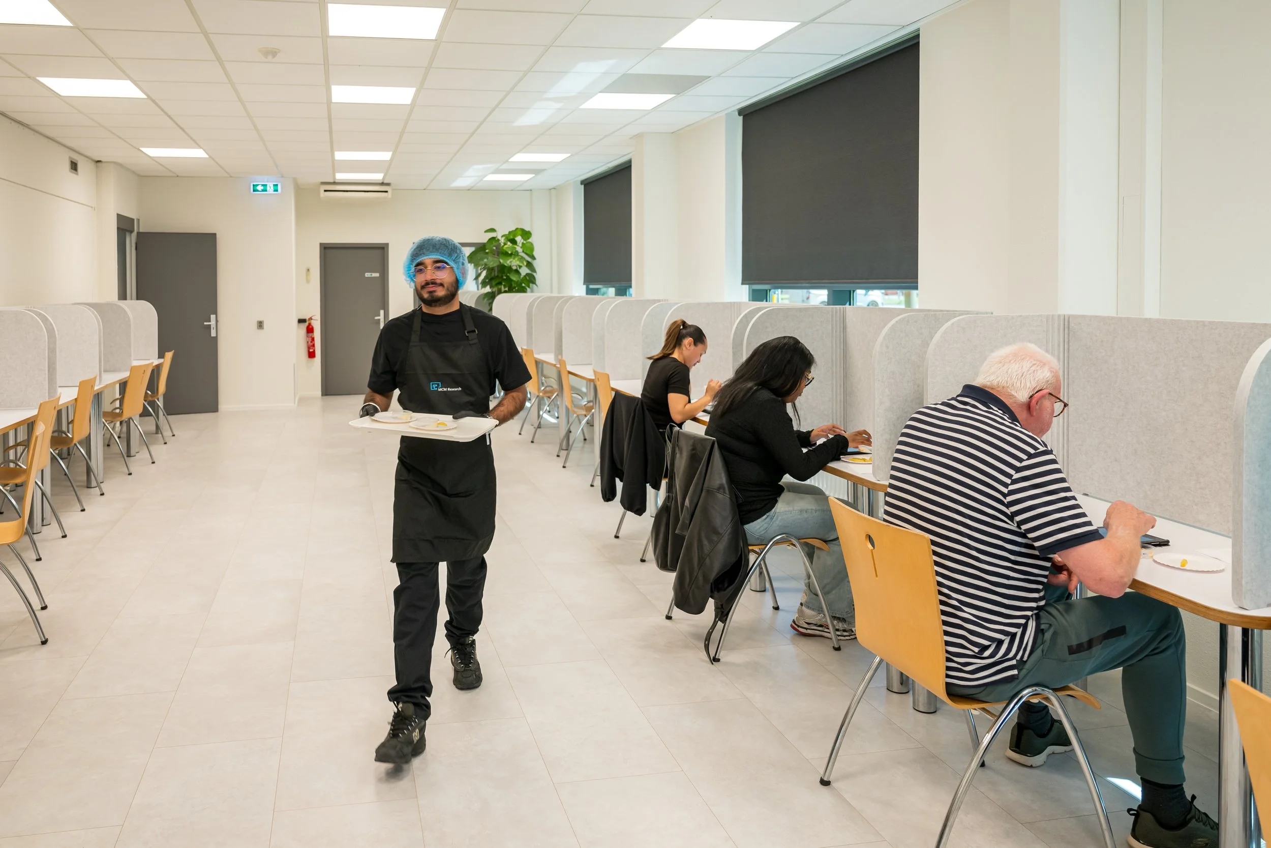 Man in zwart schort, hoed en brillen brengt een tray met eten in een restaurant. Mensen zitten gescheiden door scheidingswanden aan tafels, eten en lezen.