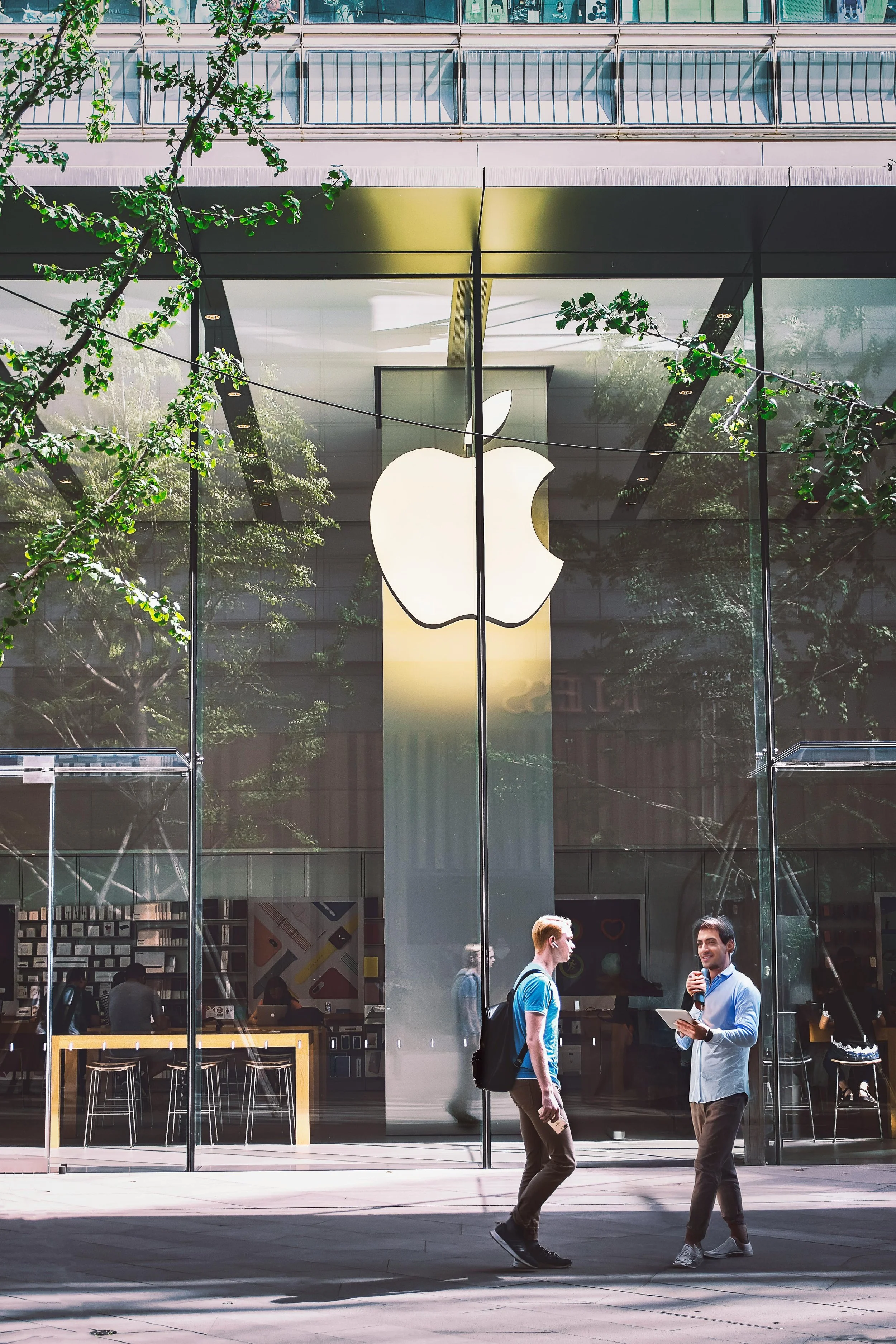 Twee mannen staan buiten in gesprek voor een Apple Store met grote ramen en zichtbaar Apple-logo. Binnen kunnen mensen werken en shoppen.