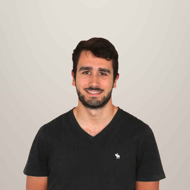 Portret van een man met donker haar en baard, gekleed in een zwart T-shirt met een klein logo, lachend voor een neutrale achtergrond.