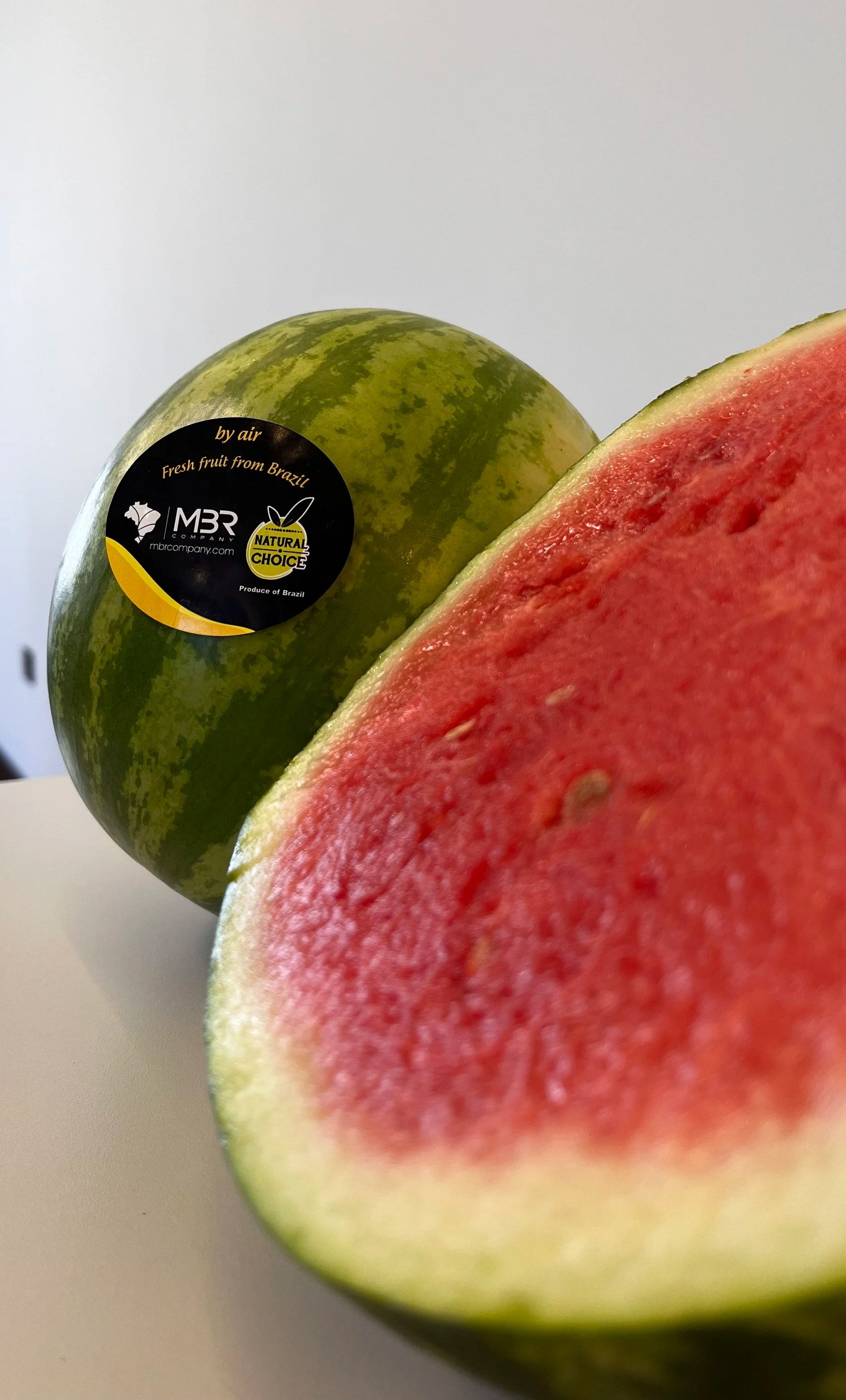 Imagem de uma melancia cortada ao meio, mostrando sua polpa vermelha e suculenta, ao lado de uma melancia inteira com etiqueta que indica ser uma fruta fresca do Brasil.