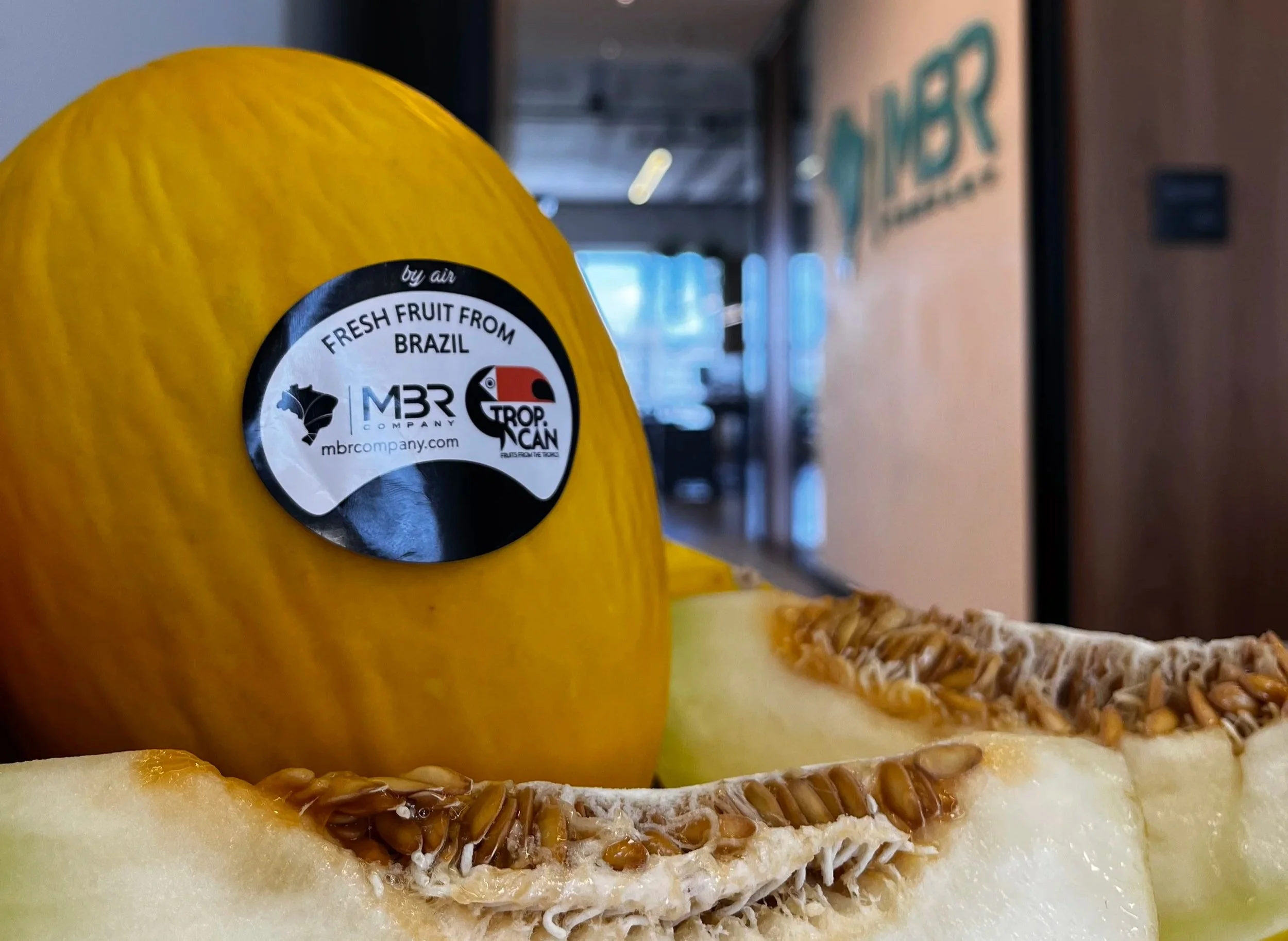 Maçã amarelo com uma etiqueta sobre ela que diz 'Fruta fresca do Brasil', ao fundo uma prateleira de supermercado com produtos.