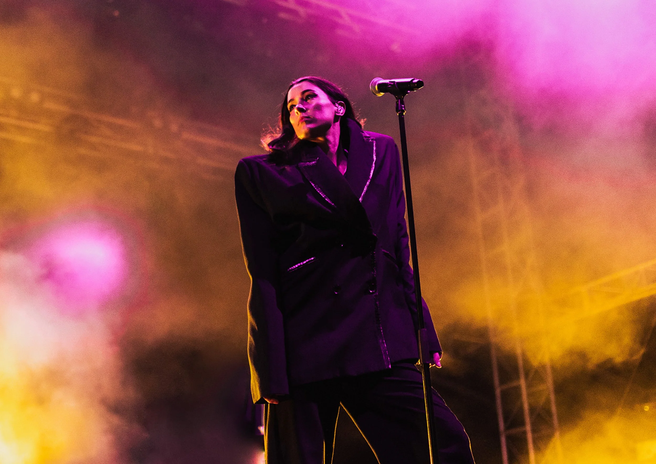 A female singer in a dark jacket performs on stage surrounded by colorful stage lighting.