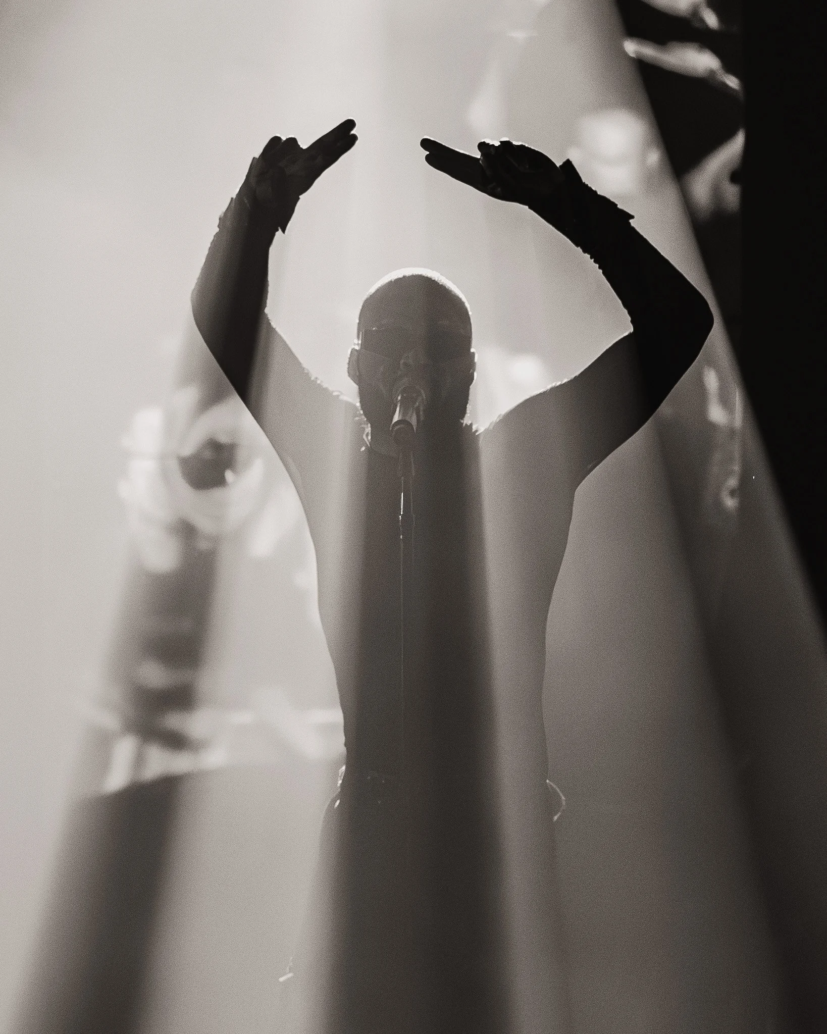 A performer with glasses and a microphone on stage, silhouetted against bright lights, raising their hands.