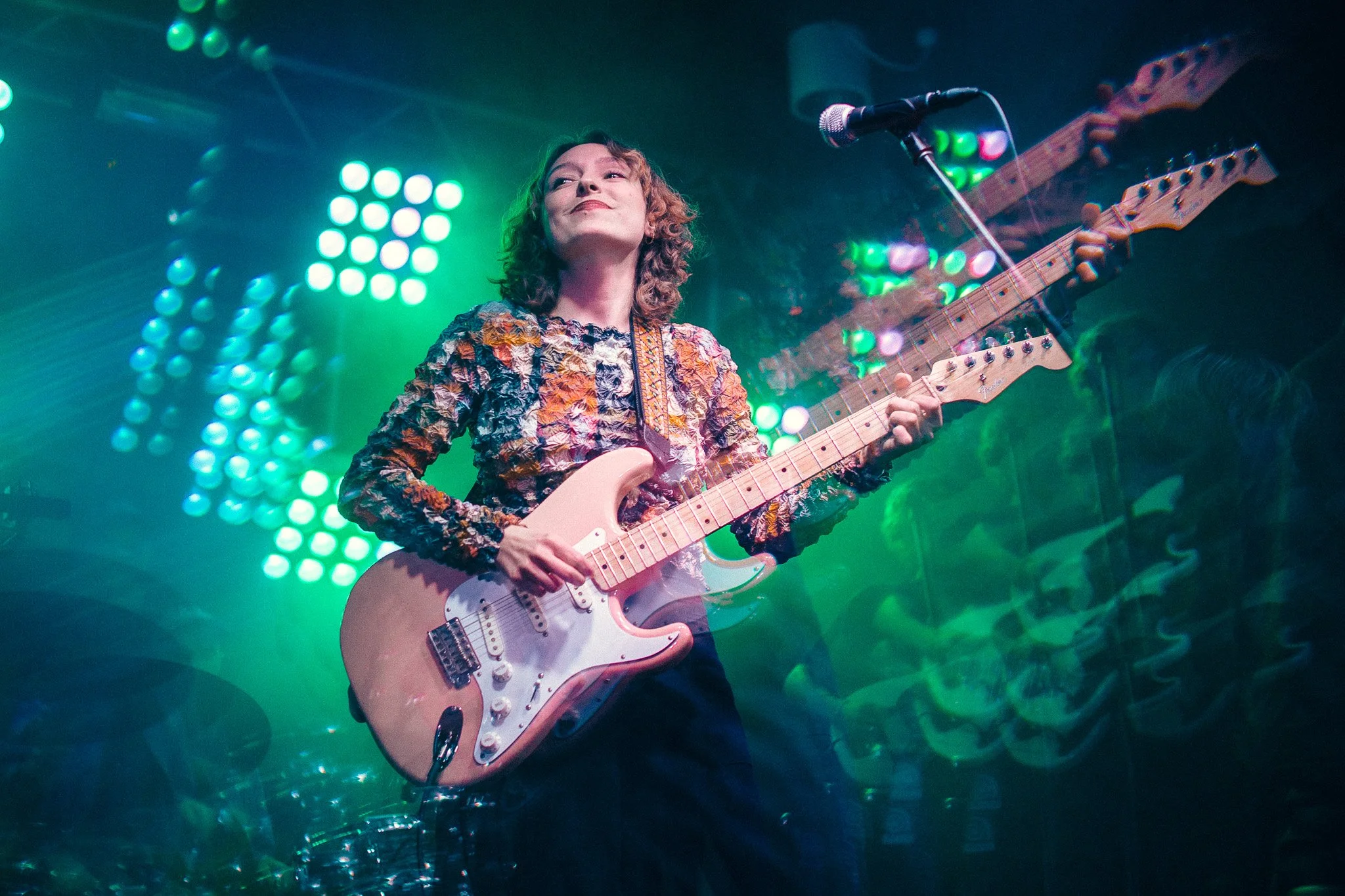 A woman with curly hair playing an electric guitar on stage under colorful, vibrant lights.