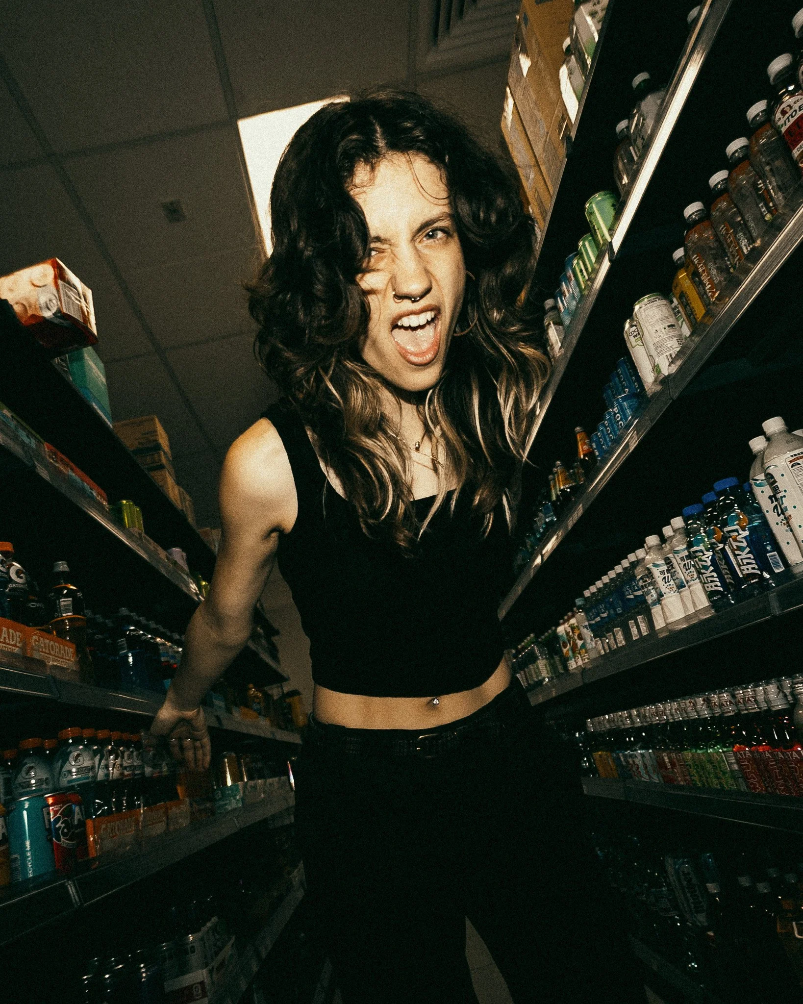 A woman with black hair, wearing a black top and a navel ring, standing between store shelves filled with canned and bottled beverages in a store aisle, making a facial expression with her mouth open and one eye closed.