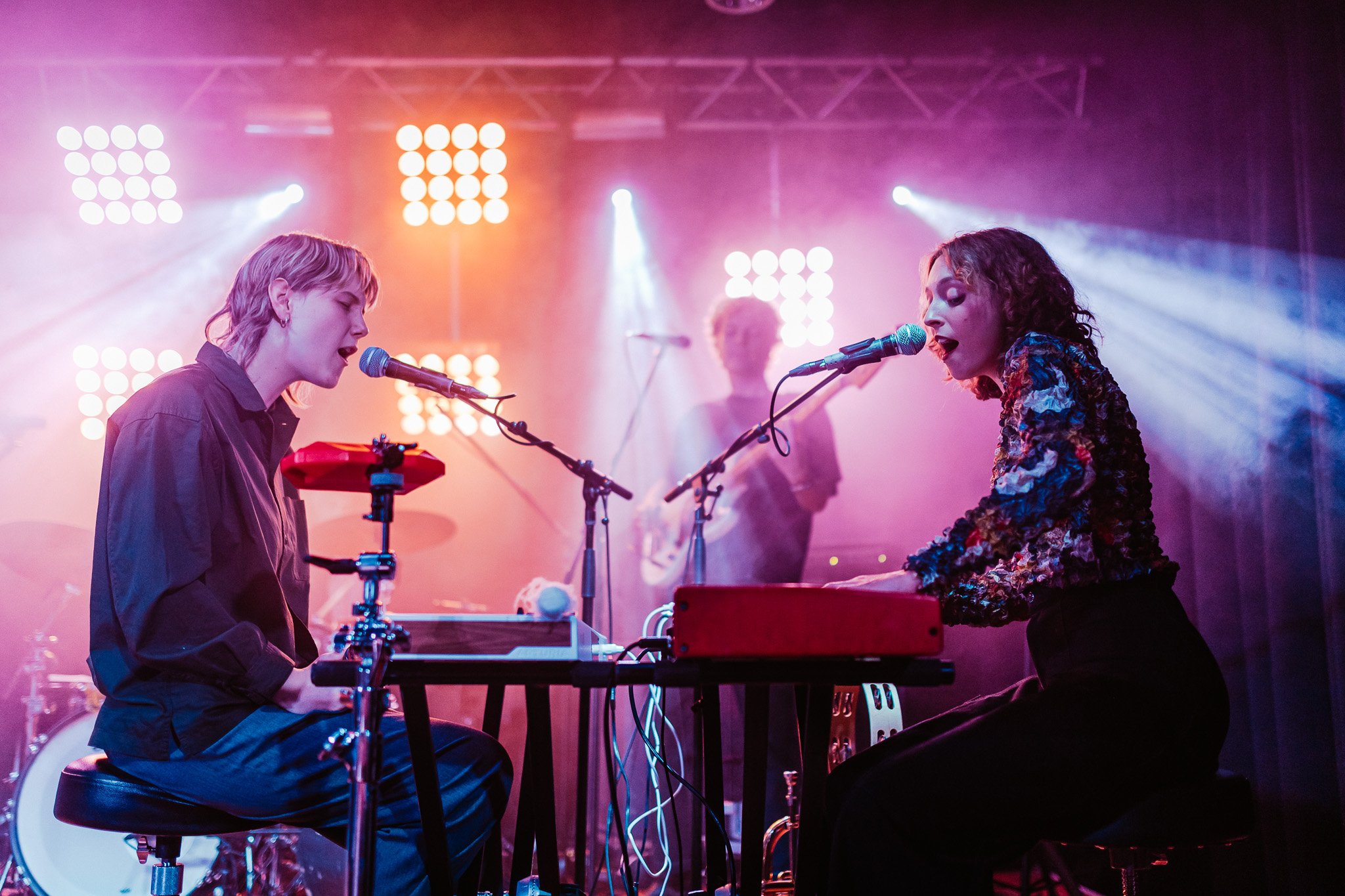 Two women are playing keyboards and singing into microphones during a live music performance on stage. They are surrounded by colorful stage lights with a band in the background.