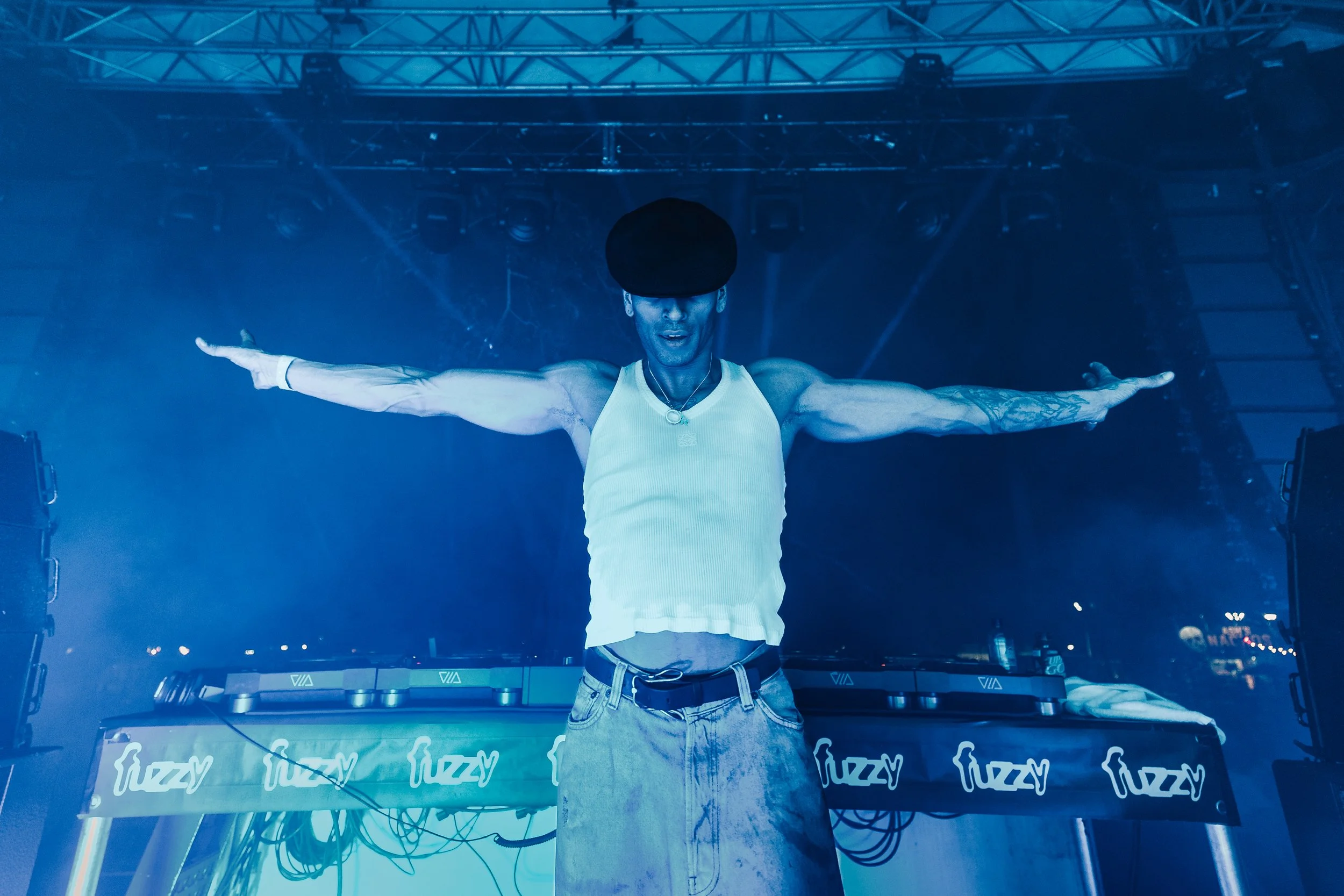 A person with tattoos on their arms wears a yellow tank top, a large black hat, and light blue jeans, standing with arms outstretched on a stage surrounded by stage equipment and blue lighting.
