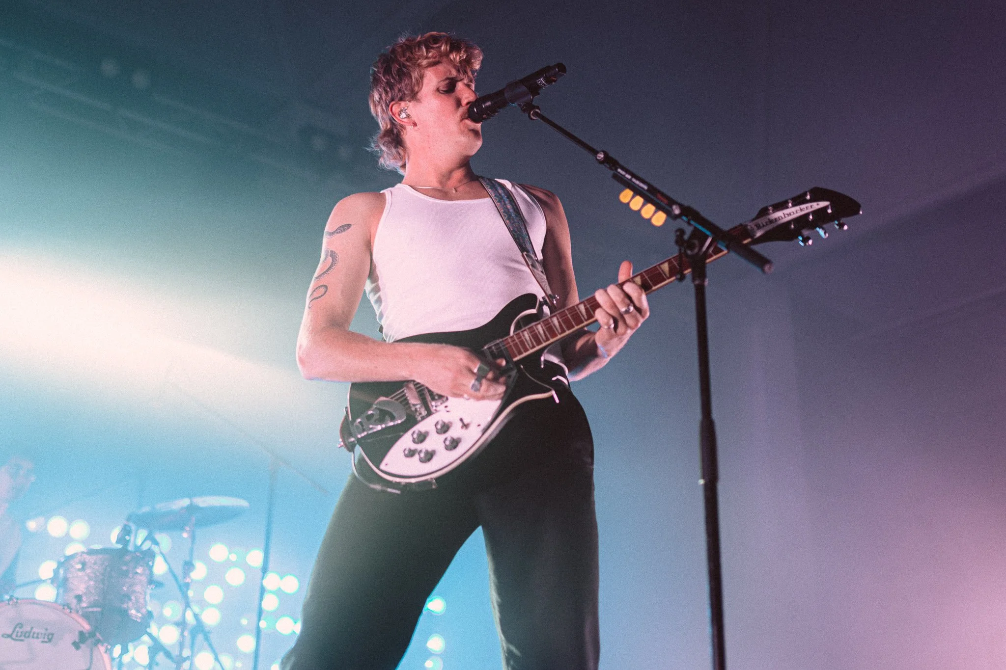 A woman with short curly hair playing an electric guitar and singing into a microphone on stage with colorful lighting.