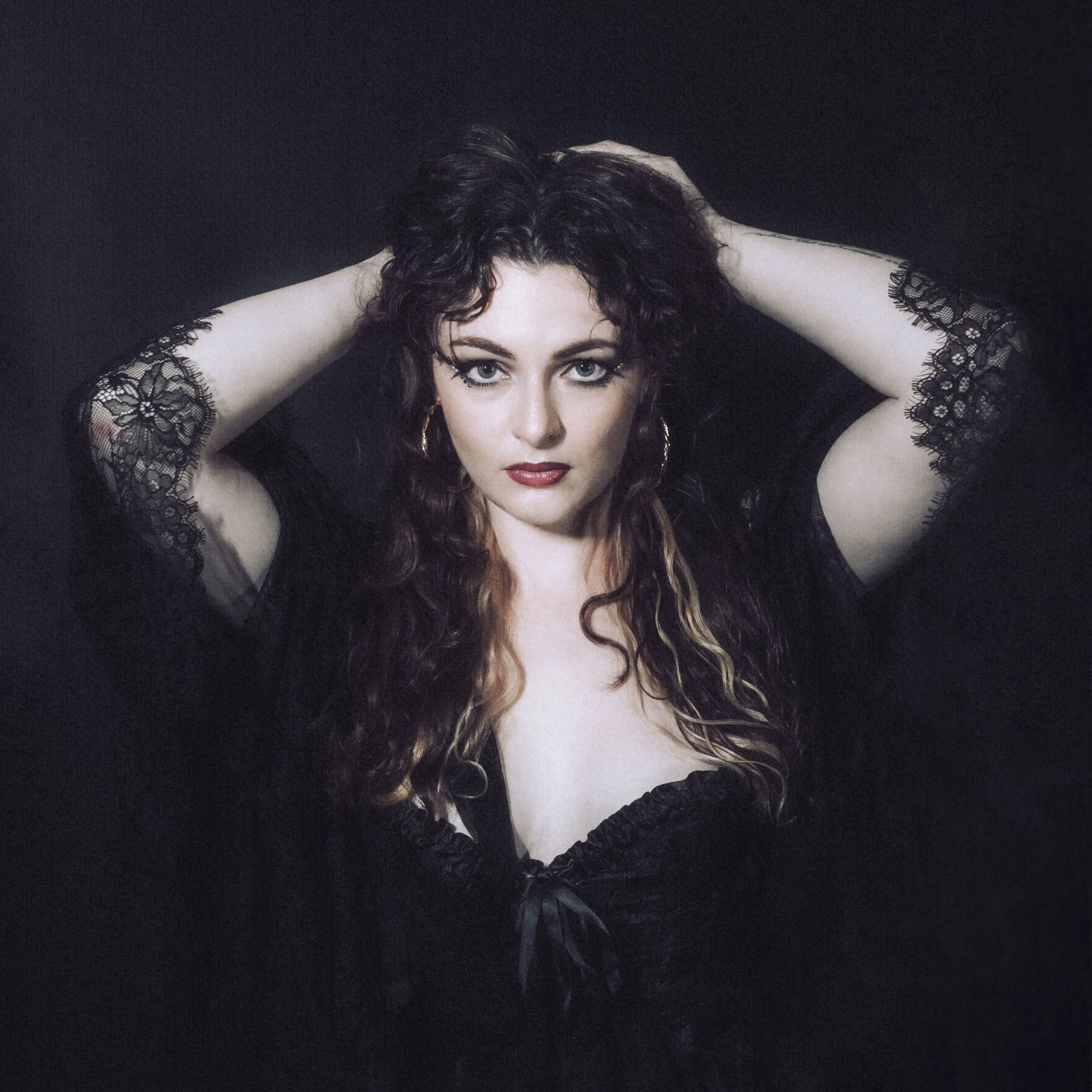 A woman with curly brown hair and striking eye makeup posing with her hands on her head against a dark background, wearing a black lace top.