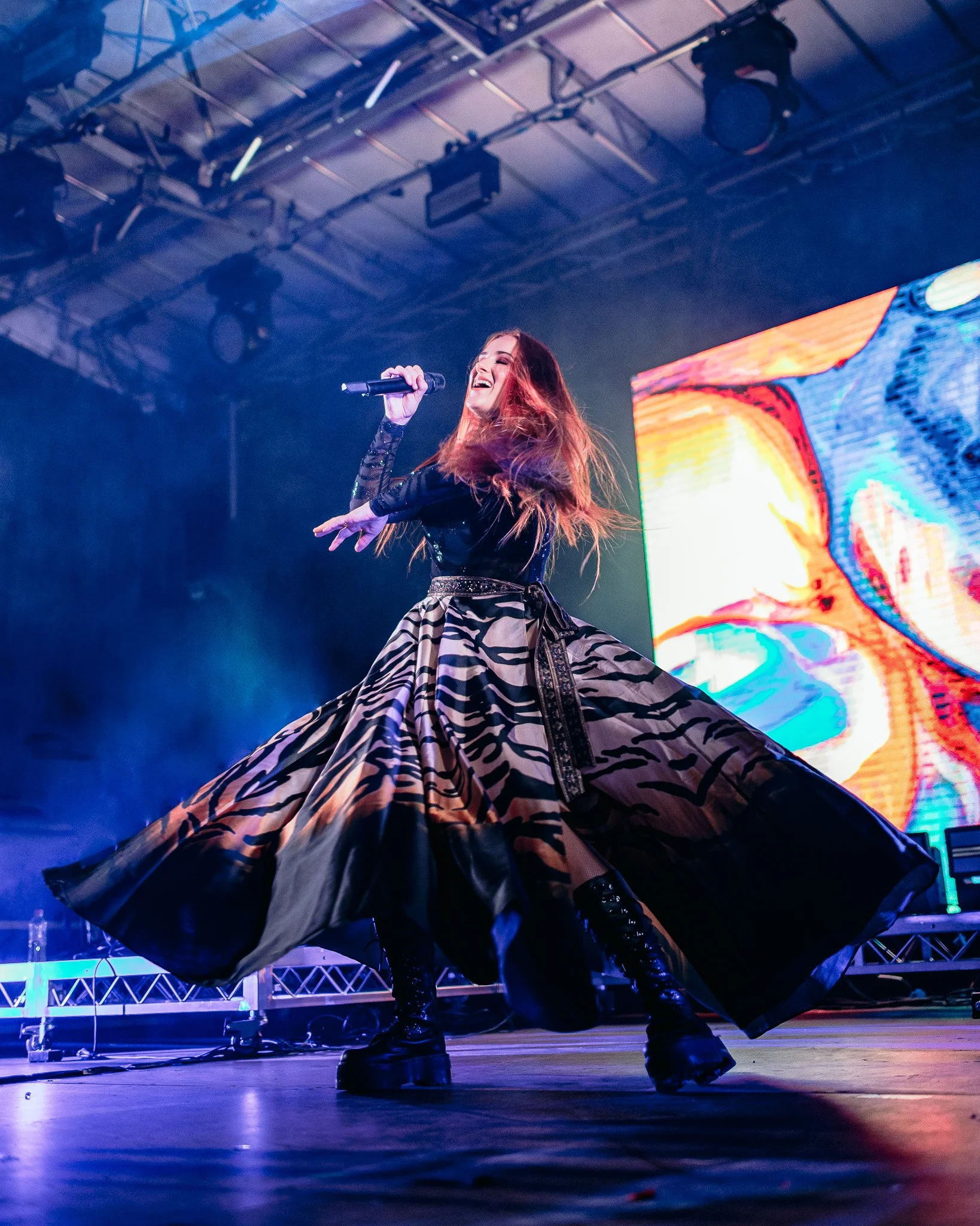 Performer on stage with long red hair, wearing a black top with long sleeves, a patterned skirt, and shiny black boots, dancing and holding a microphone in a large indoor concert venue with colorful digital screens in the background.