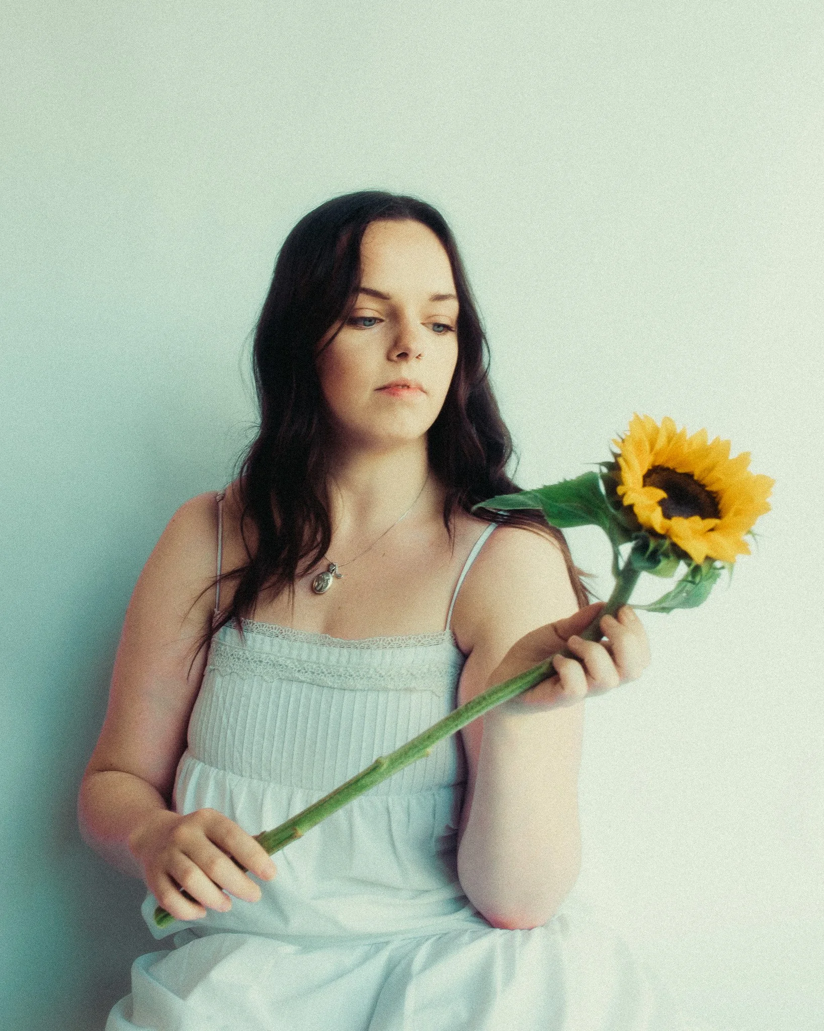 A woman with dark hair and blue eyes holding a sunflower against a light-colored wall.