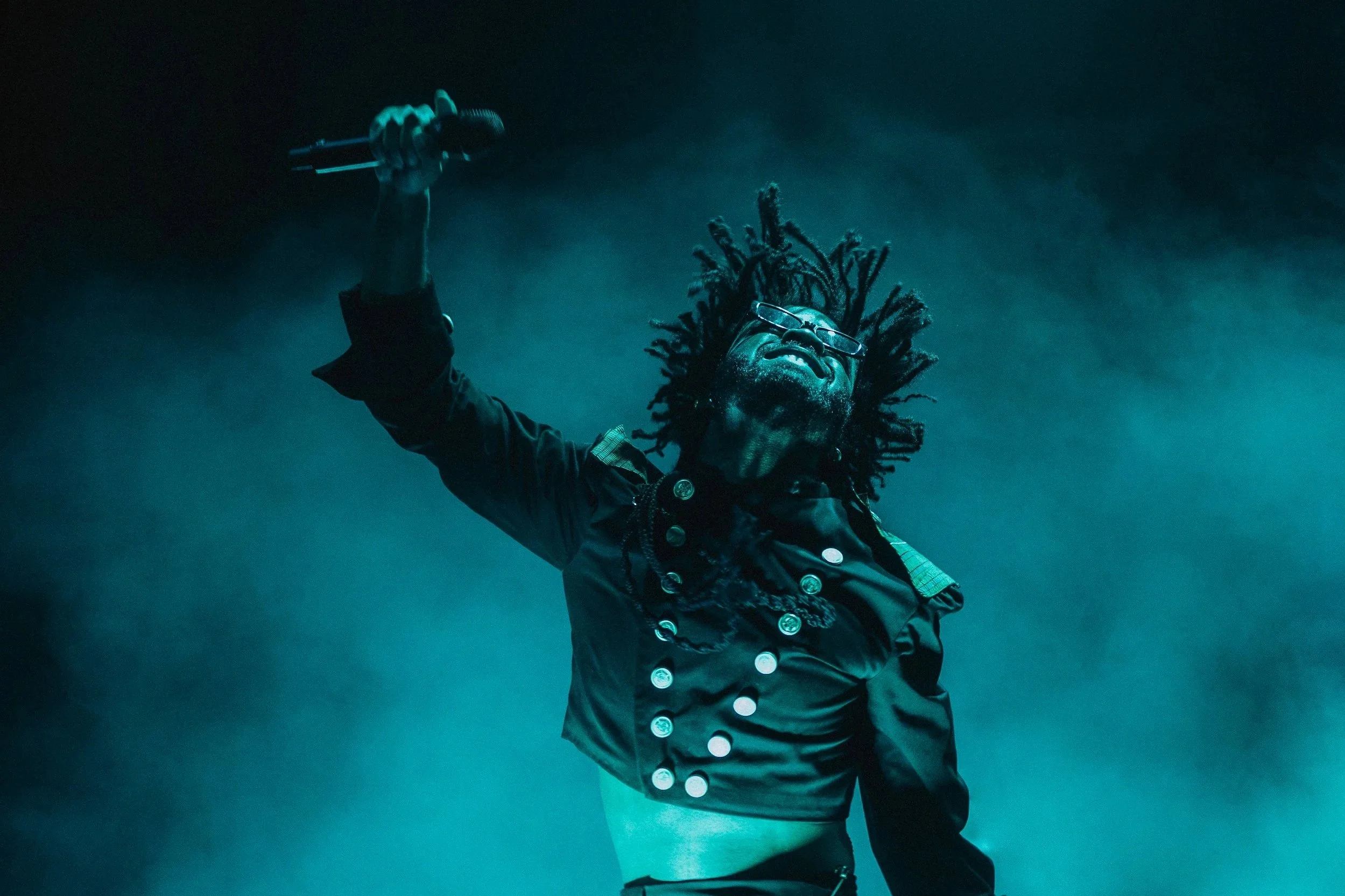 A person with dreadlocks is on stage, raising a microphone in one hand, dressed in a military-style jacket with badges, in a dark, smoky environment.