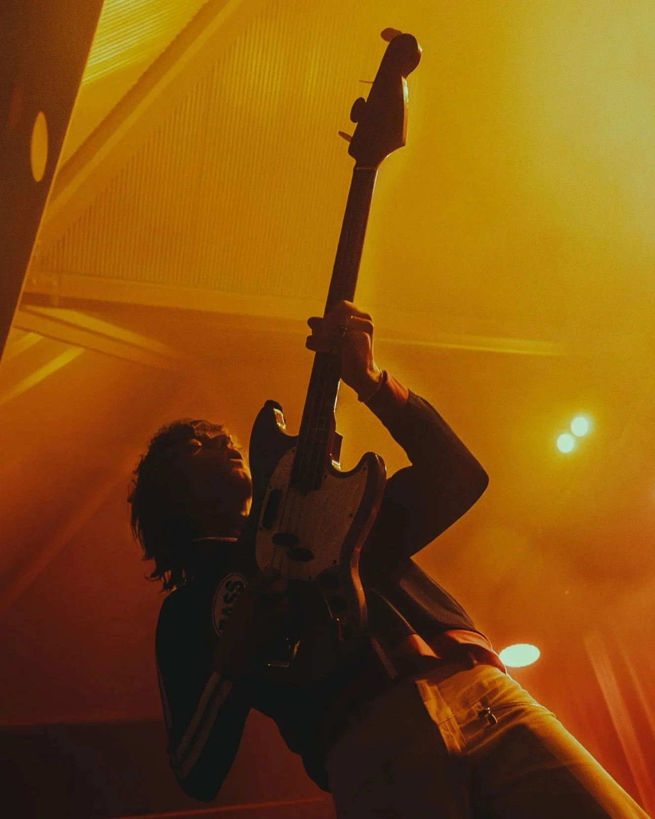 A silhouette of a person playing an electric guitar on stage, illuminated by warm yellow and orange stage lights, with a background that looks like a venue with a dome ceiling.