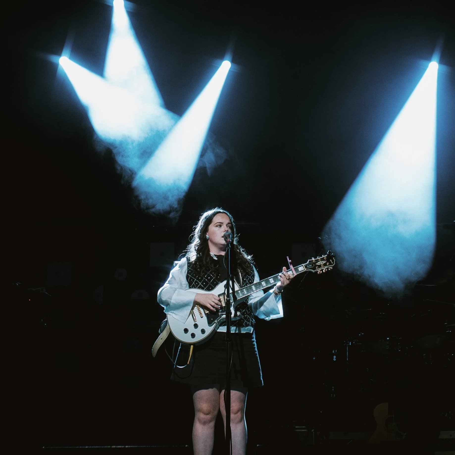 A woman with long dark hair singing into a microphone while playing a white electric guitar on a dark stage with bright spotlights shining down.