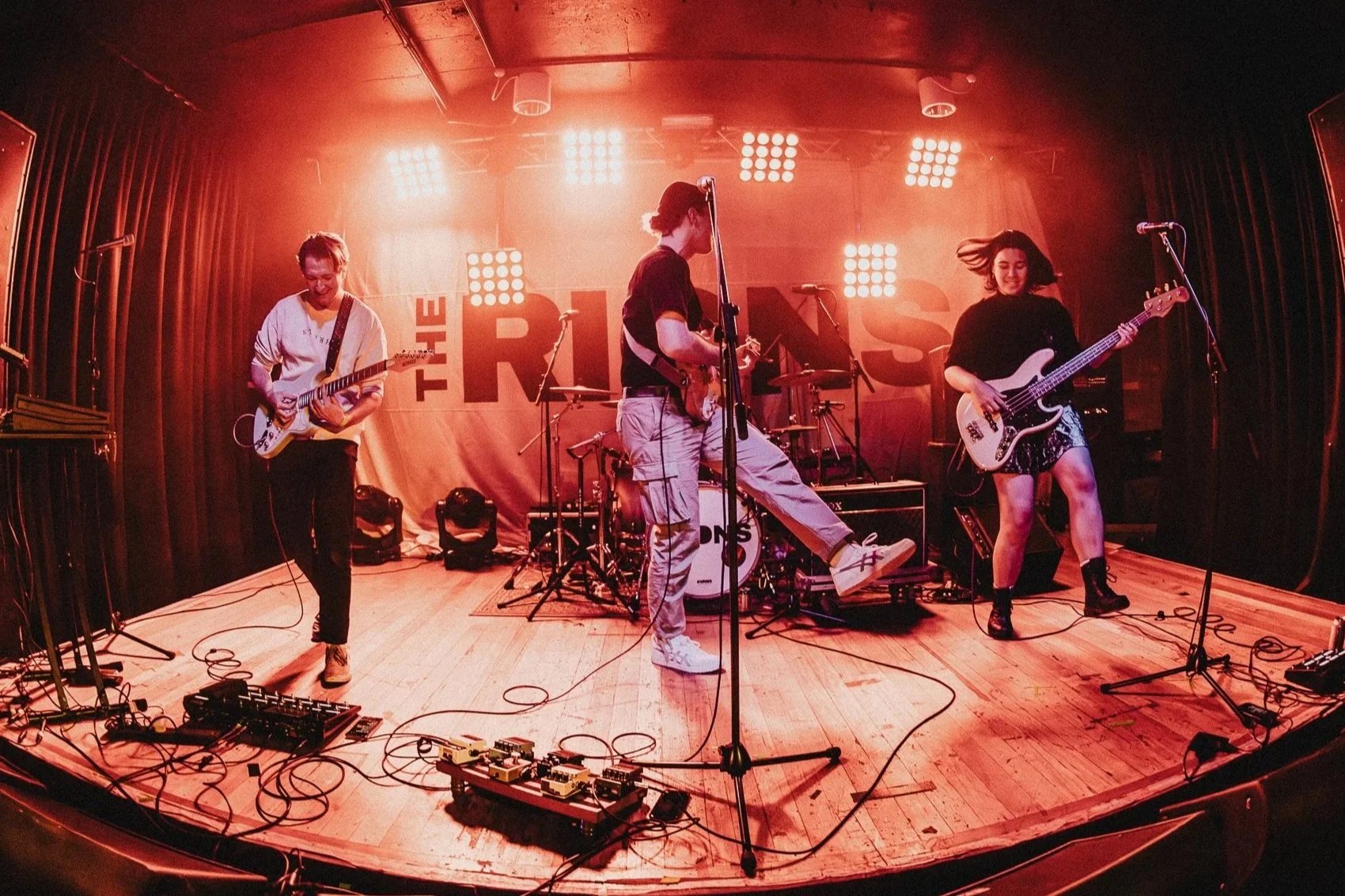 A band performing on a stage with red lighting, with three musicians playing guitars and a drummer in the background, and equipment including pedals and microphones in the foreground.