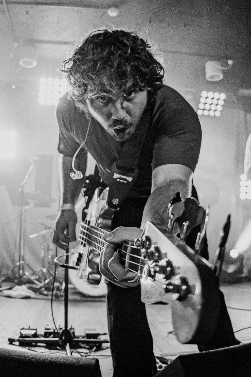 A male musician with curly hair passionately playing an electric guitar on stage during a live performance, with stage lights in the background.