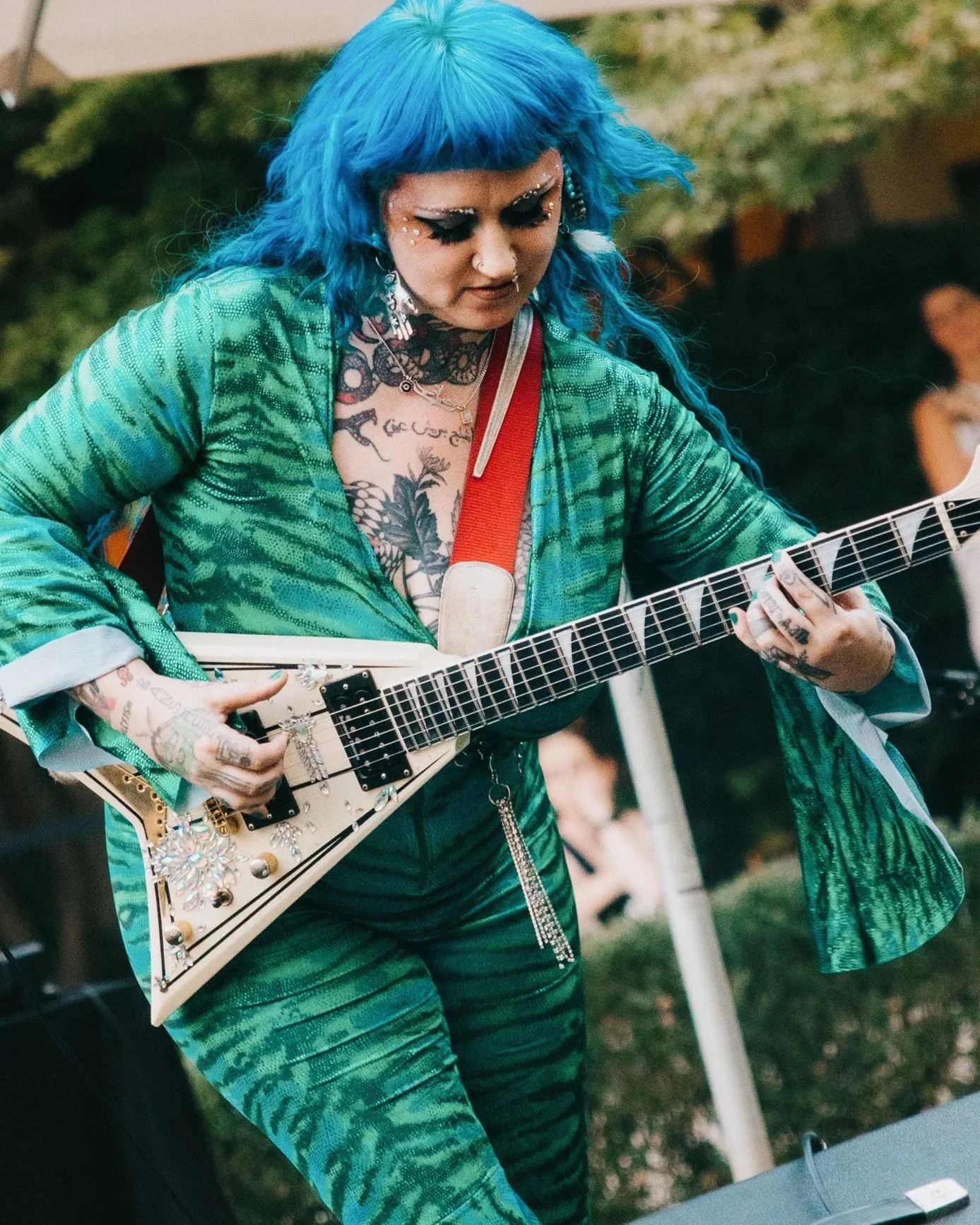 A woman with bright blue hair and tattoos playing a white electric guitar decorated with rhinestones at an outdoor event.