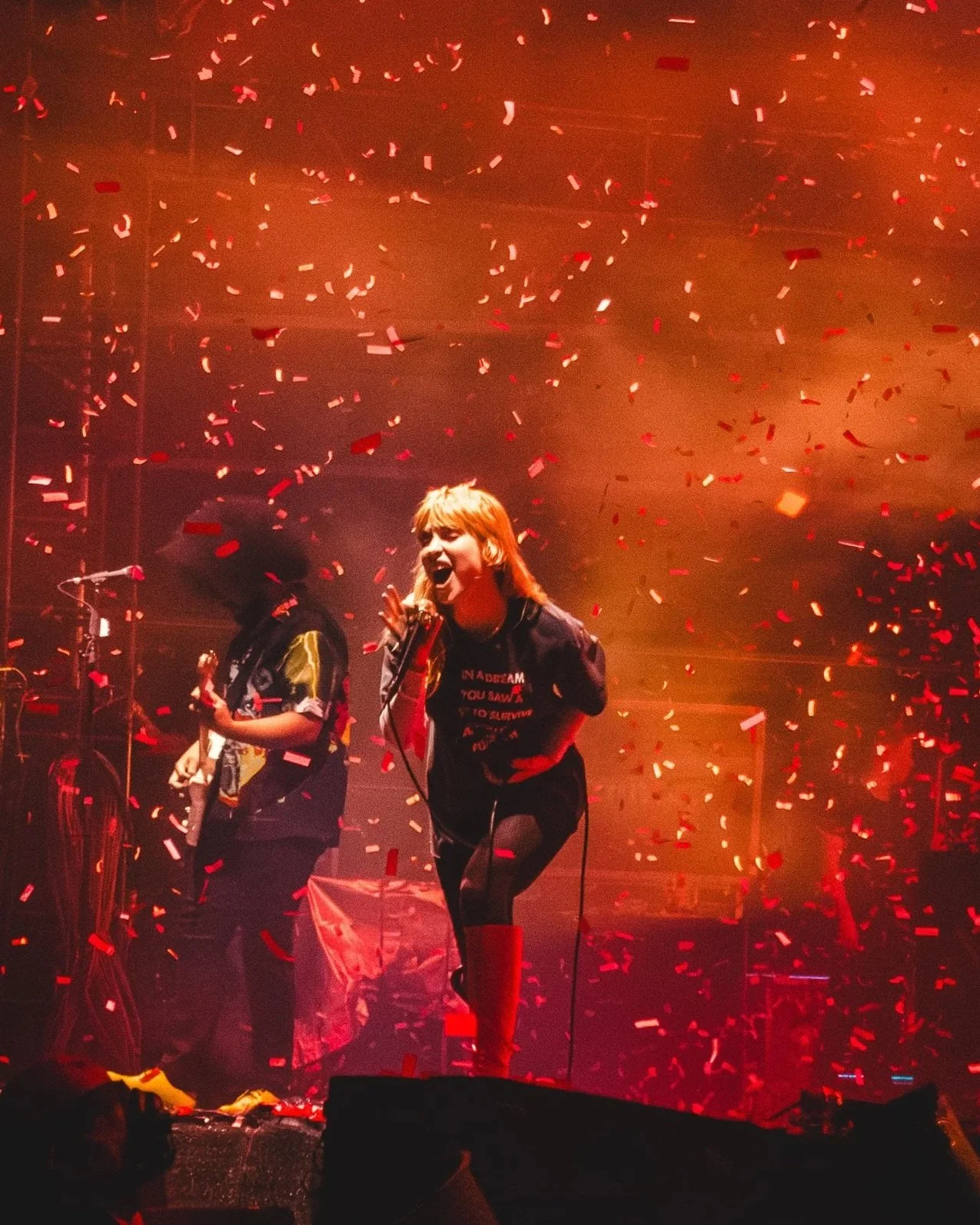 A woman singing on stage with red confetti falling around her, accompanied by a musician playing guitar in the background, with stage lighting creating a warm glow.