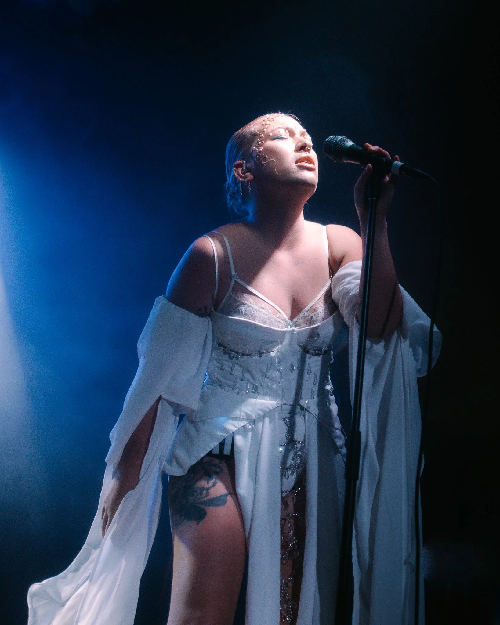 A woman singing into a microphone on stage, dressed in a sheer white outfit with sequins, with her eyes closed and head slightly tilted back, illuminated by stage lighting.