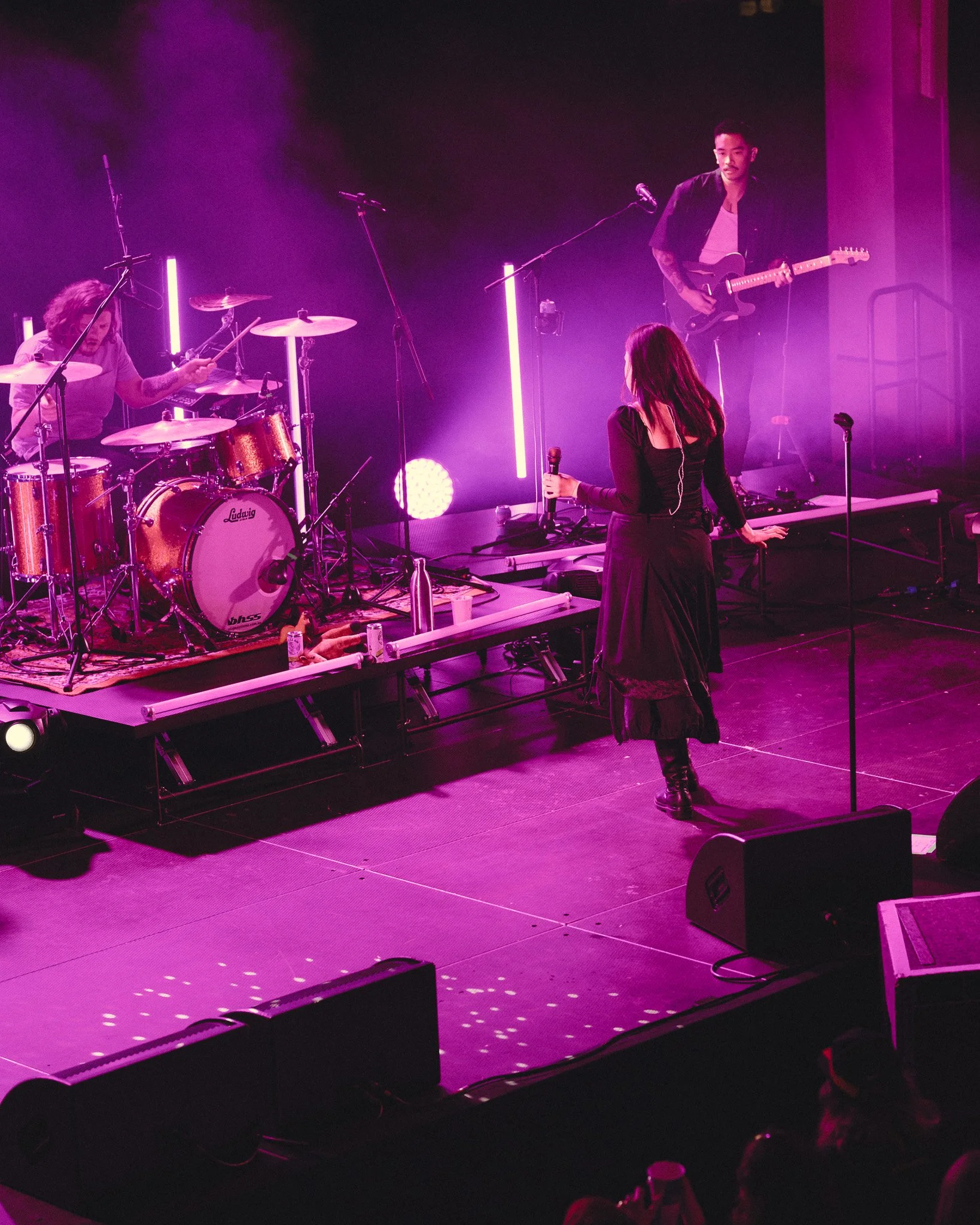 A live musical performance on stage with purple lighting, featuring a woman with long dark hair singing into a microphone, a drummer with curly hair playing a drum set, and a man with short hair and tattoos playing an electric guitar.