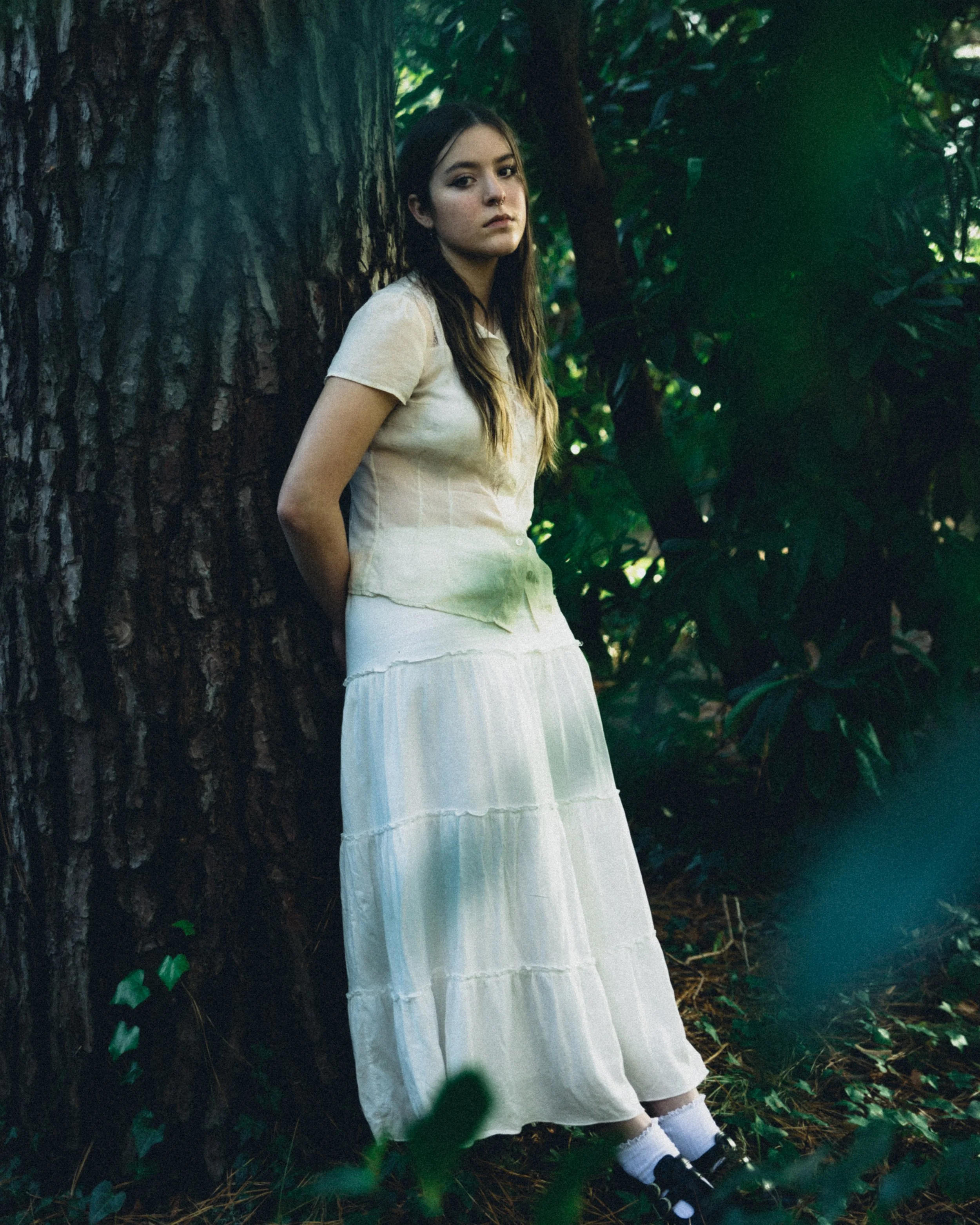 A young woman with long brown hair, wearing a white dress and black shoes with white socks, leaning against a large tree trunk in a lush green forest. She has a neutral expression and is looking off into the distance.