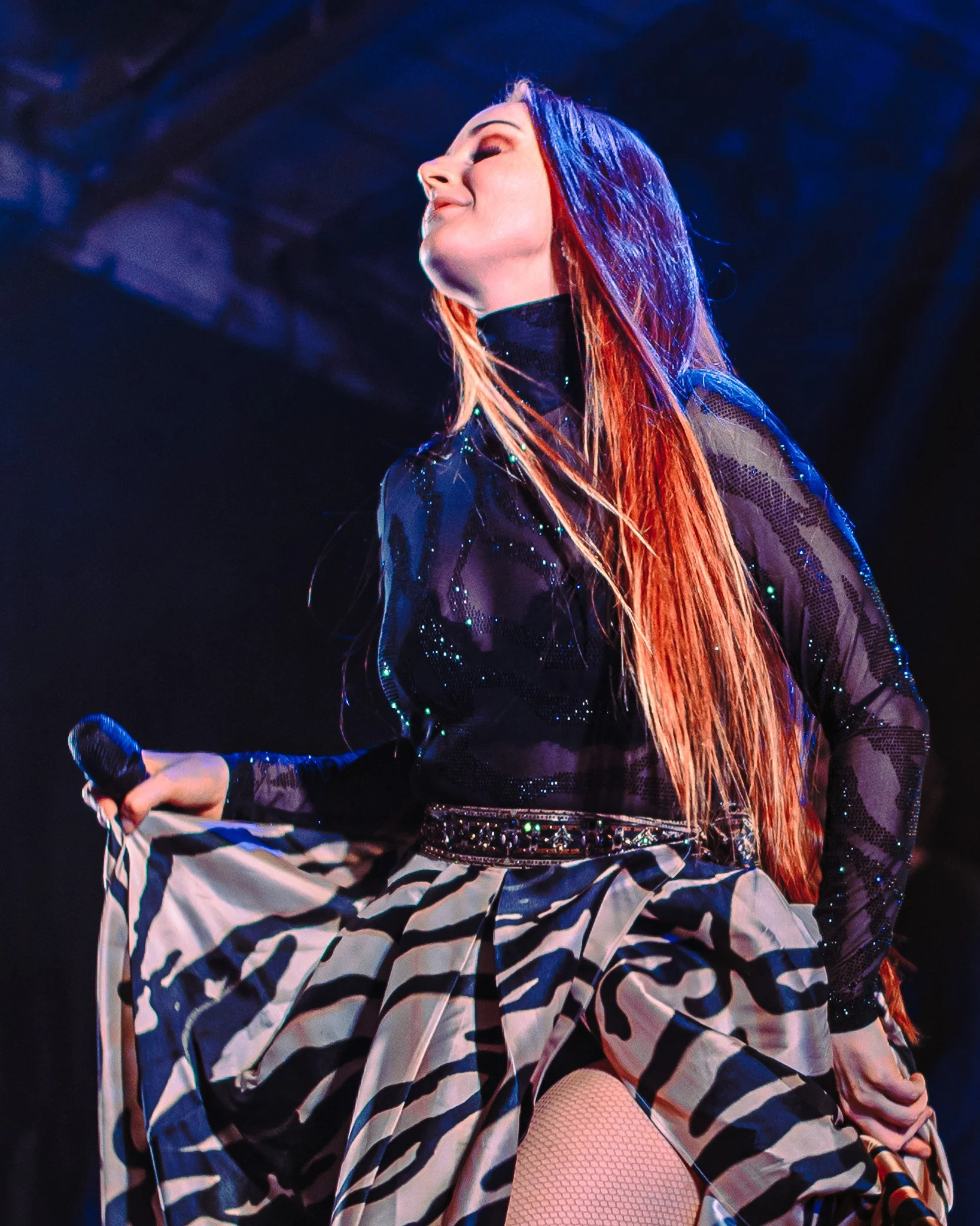 A performer on stage with long, orange hair, wearing a black sheer top with sparkles, a wide belt, striped skirt, fishnet stockings, and holding a microphone in her right hand, with her head tilted back and a confident expression.
