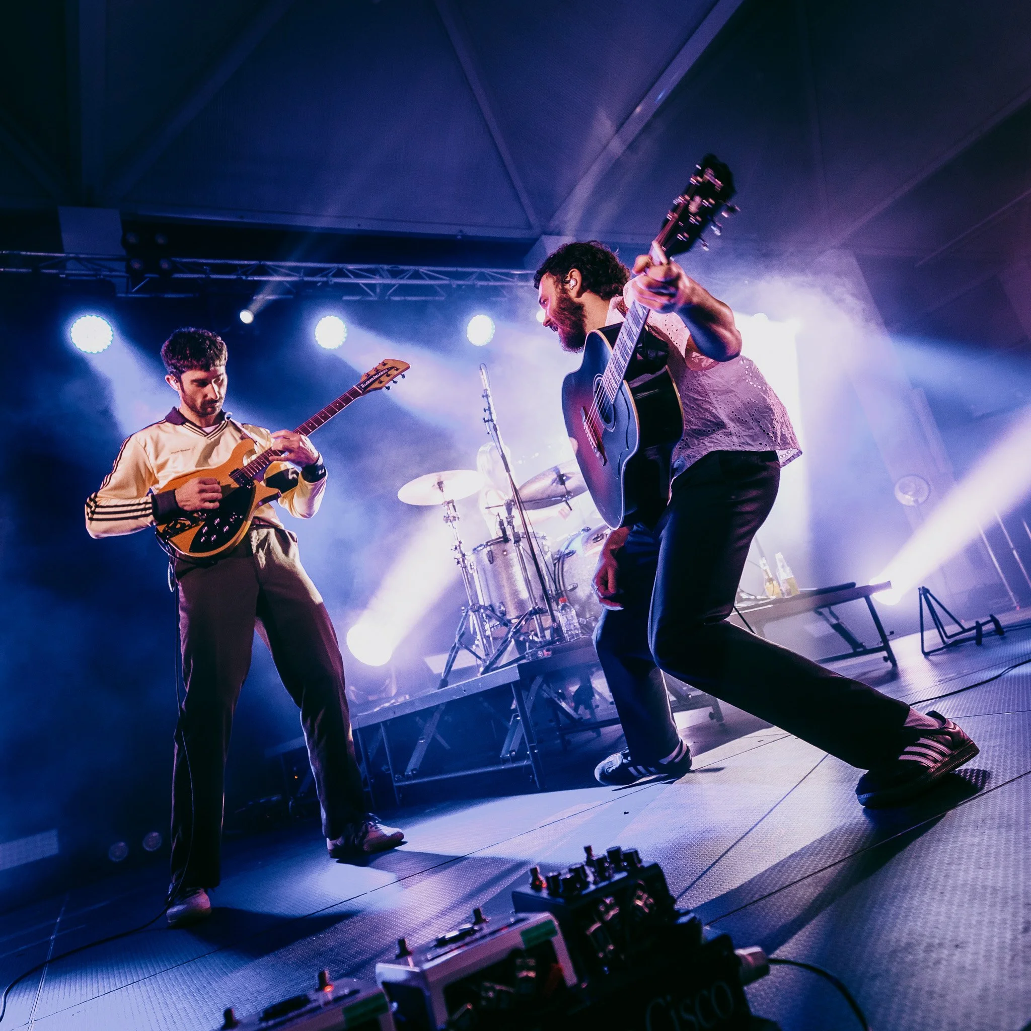Two musicians playing guitars on stage under colorful stage lights, with music equipment and a drum kit visible in the background.