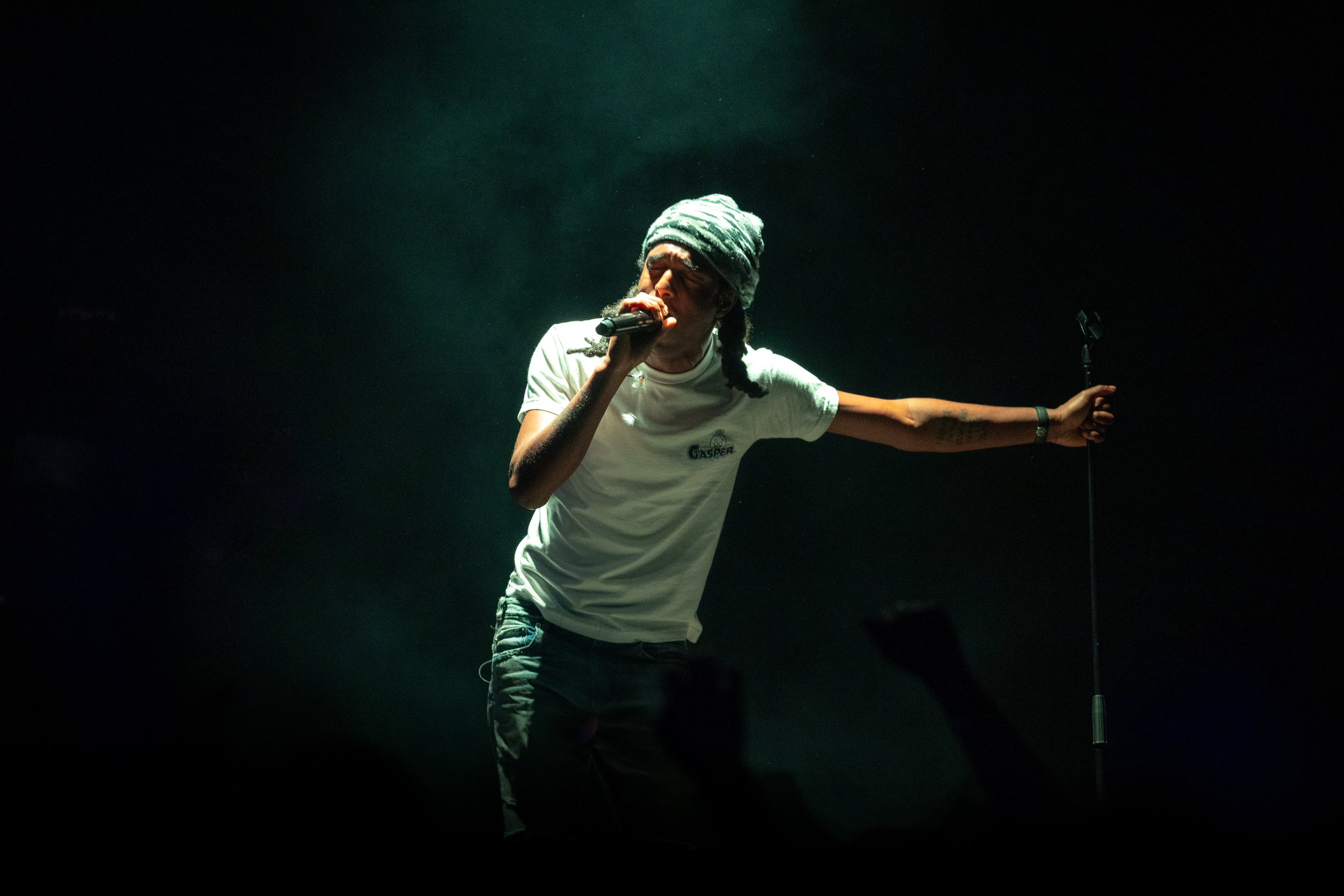 A man singing into a microphone on stage, wearing a dark beanie and a white t-shirt, with one arm stretched out holding a microphone stand, in a dark setting with focused lighting.