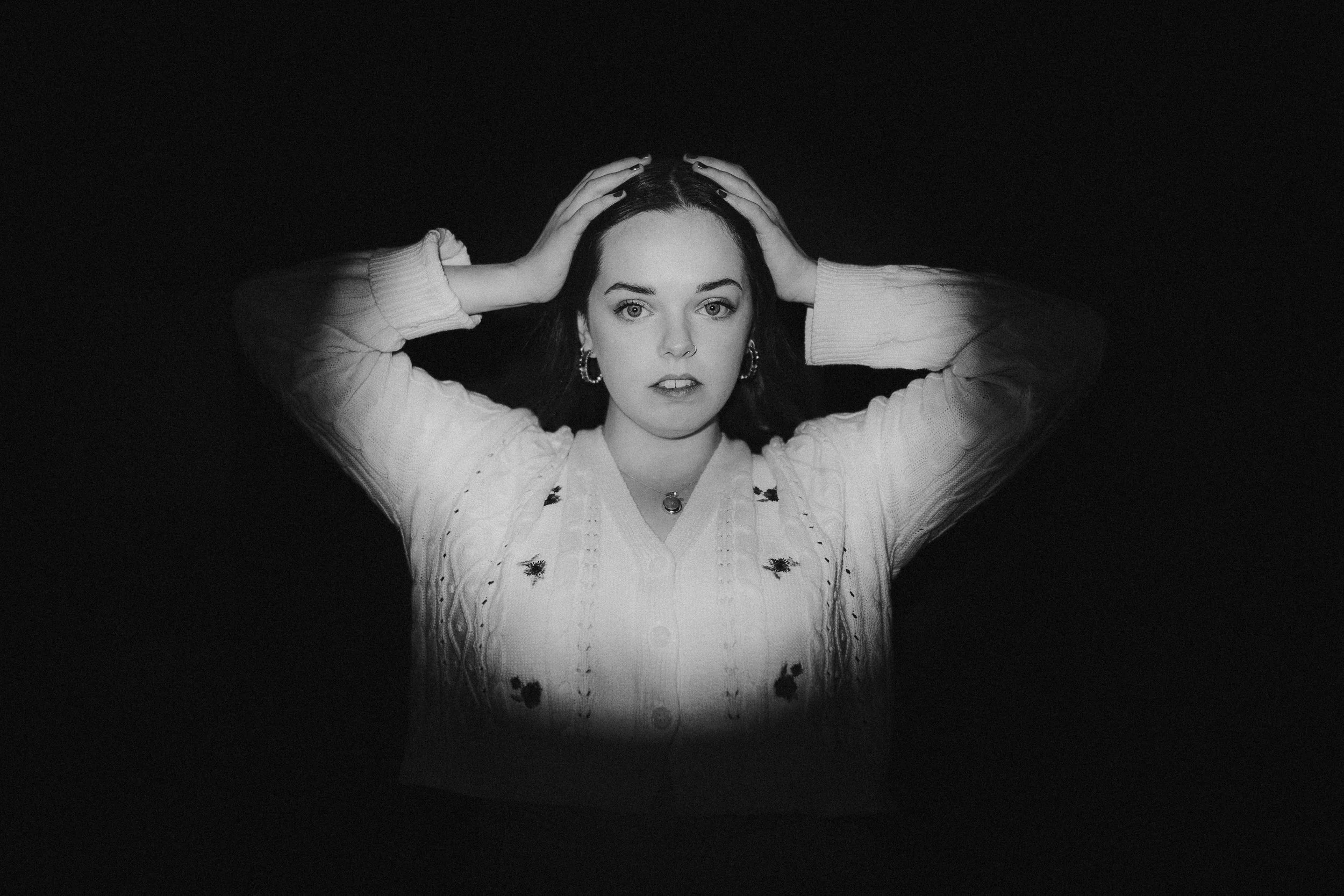 A black and white photo of a woman looking directly at the camera with her hands on her head. She has dark hair, a nose ring, earrings, and is wearing a sweater.